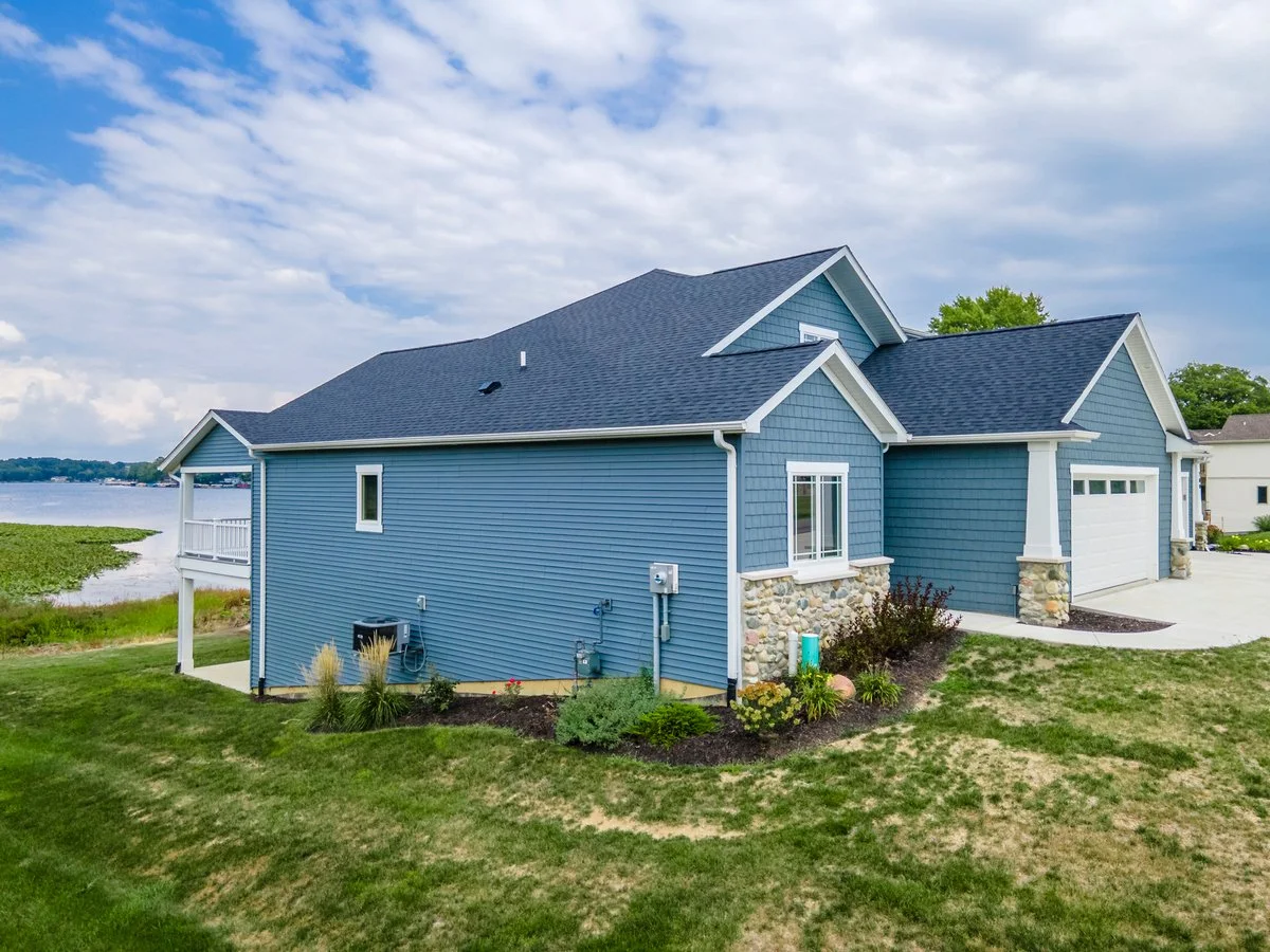 Blue house with stone accents near a body of water, with a grassy yard and clouds in the sky.