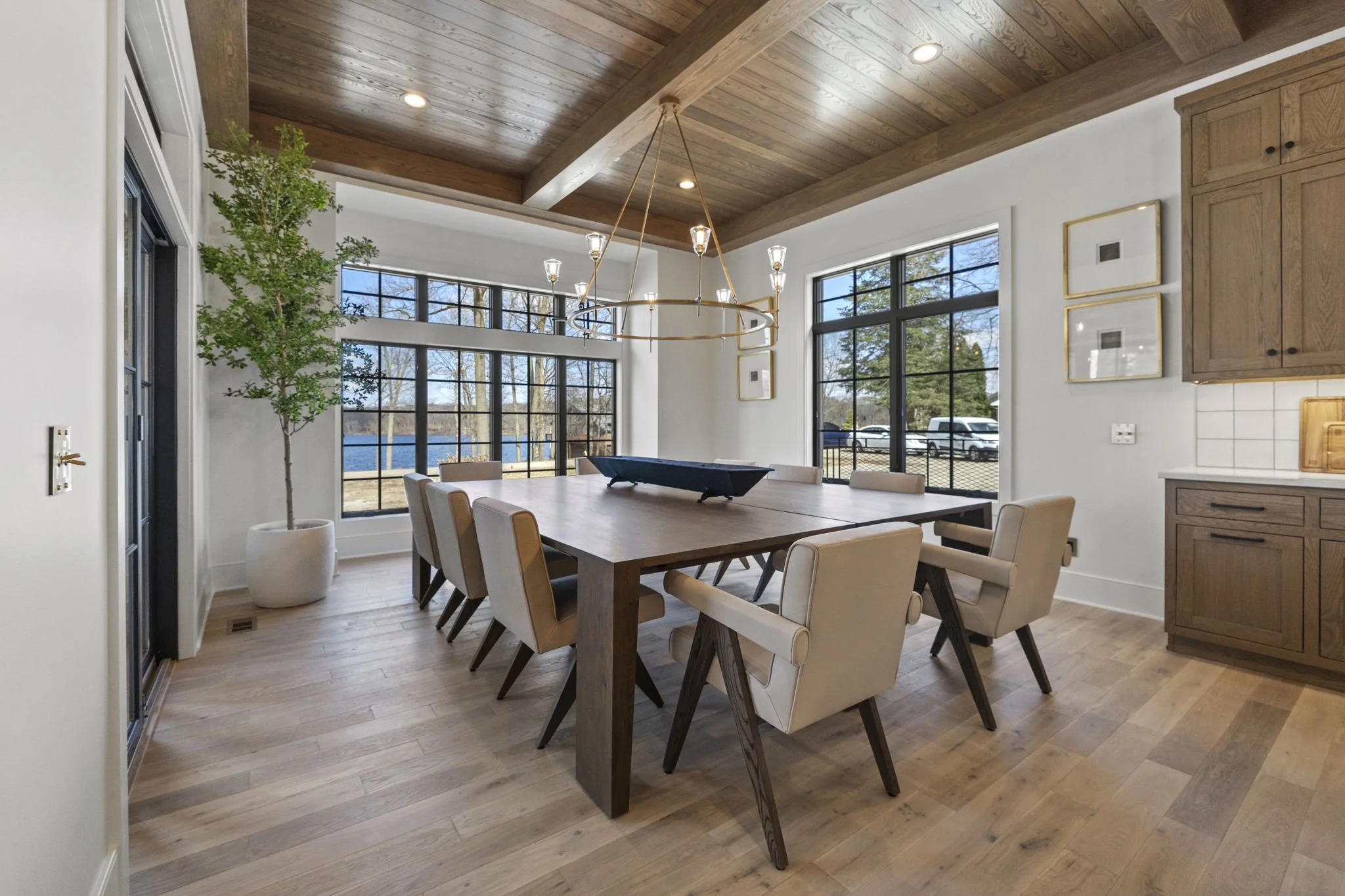 Modern dining room with large wooden table, beige chairs, a circular chandelier, wooden ceiling, large windows showing outdoors, potted plant, and some framed artwork on white walls.