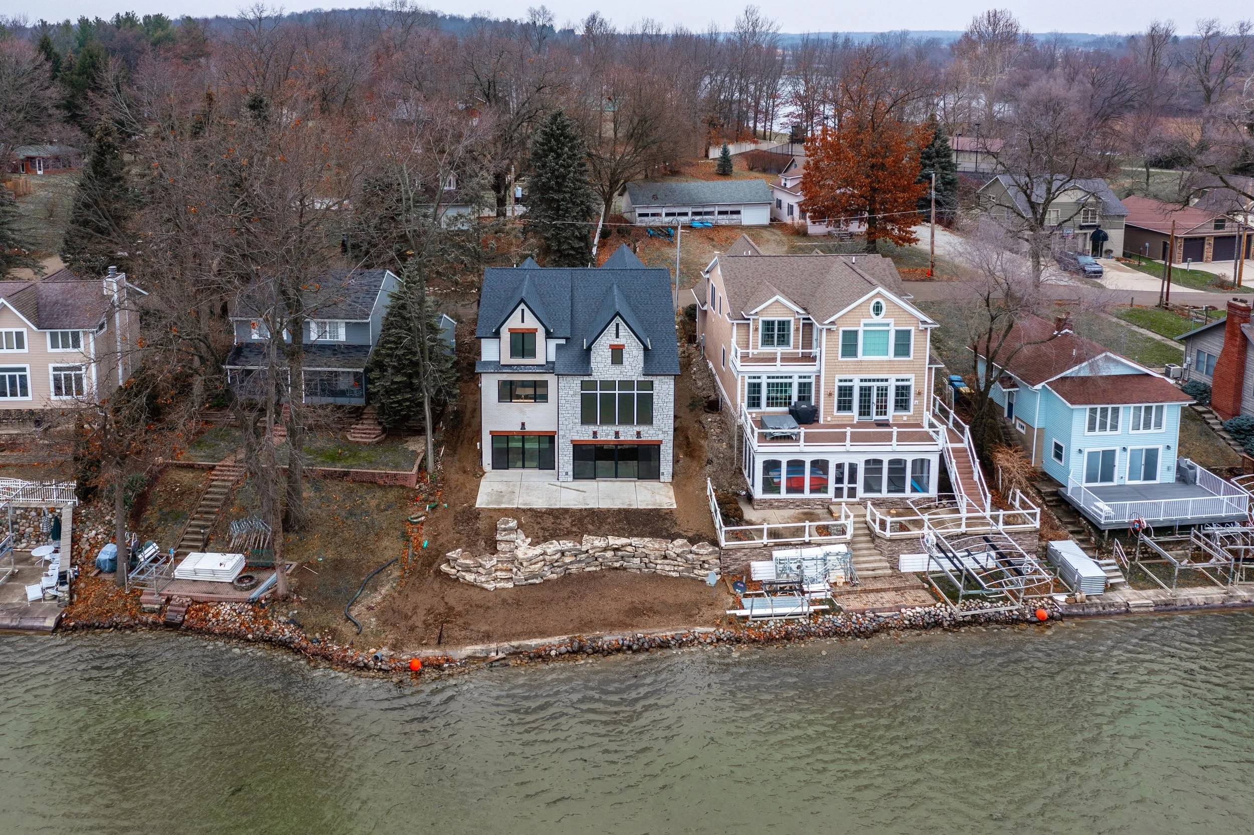 Aerial view of a row of houses along a lakeshore, with some under construction or renovation, and trees in the background.