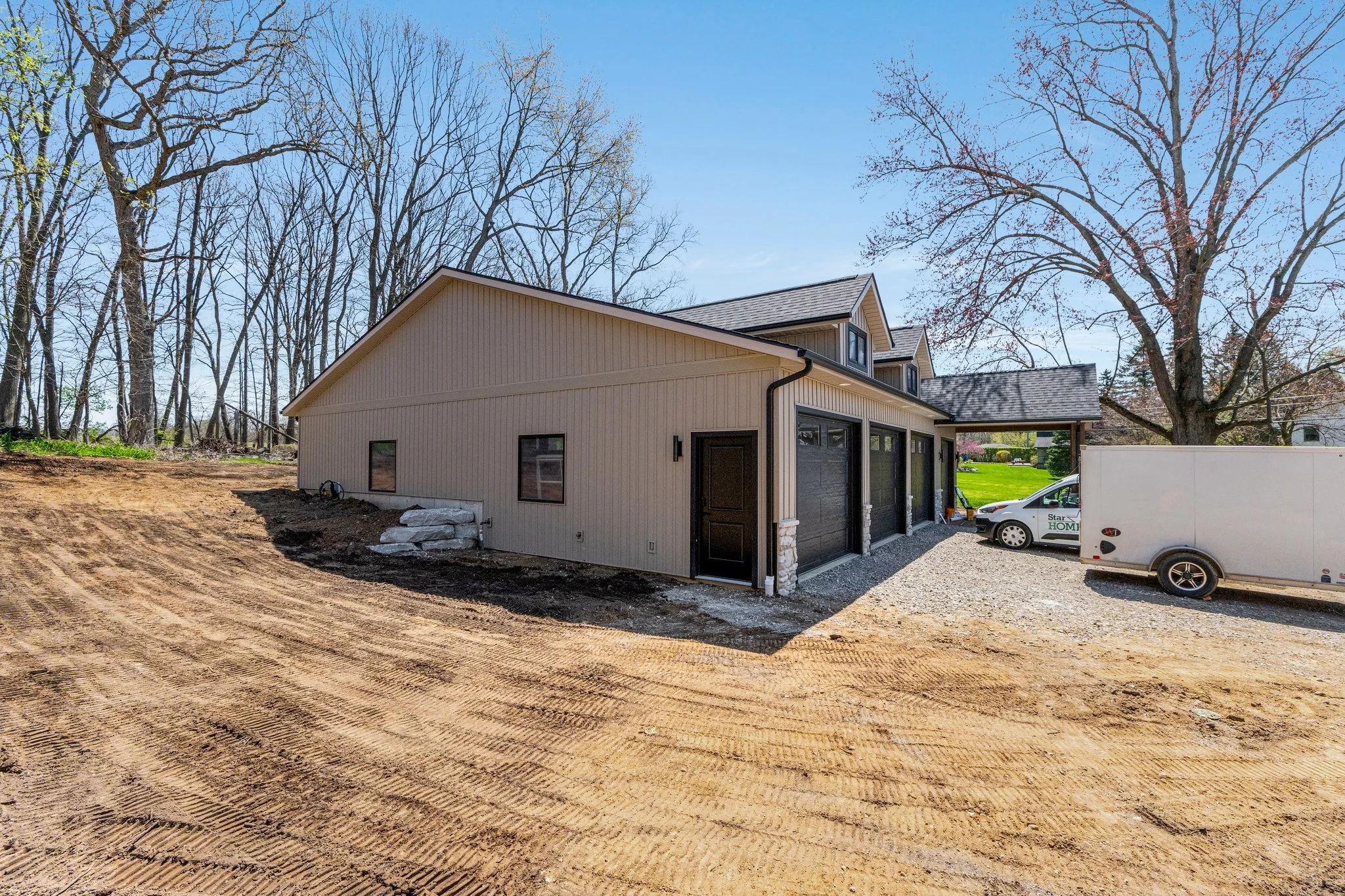 Newly constructed beige house with black garage doors, situated on a cleared lot with dirt ground, neighboring trees, and a bright blue sky.