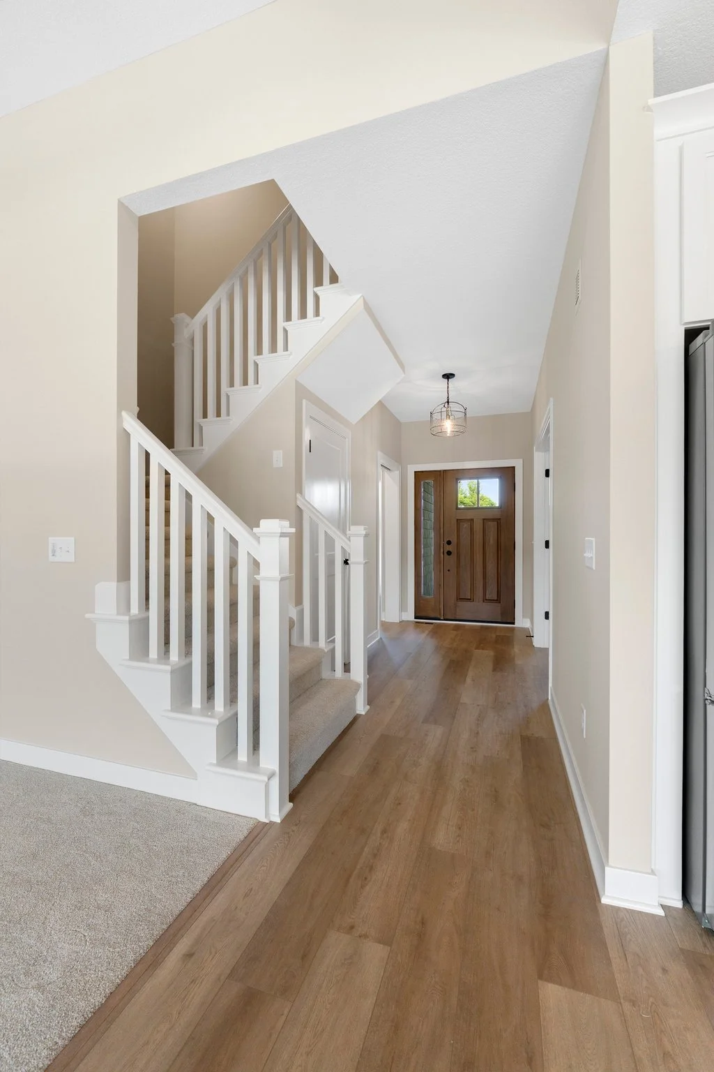 View of a bright, modern entryway with a wooden front door, hardwood flooring, staircase with white railing, beige walls, and a ceiling light fixture.
