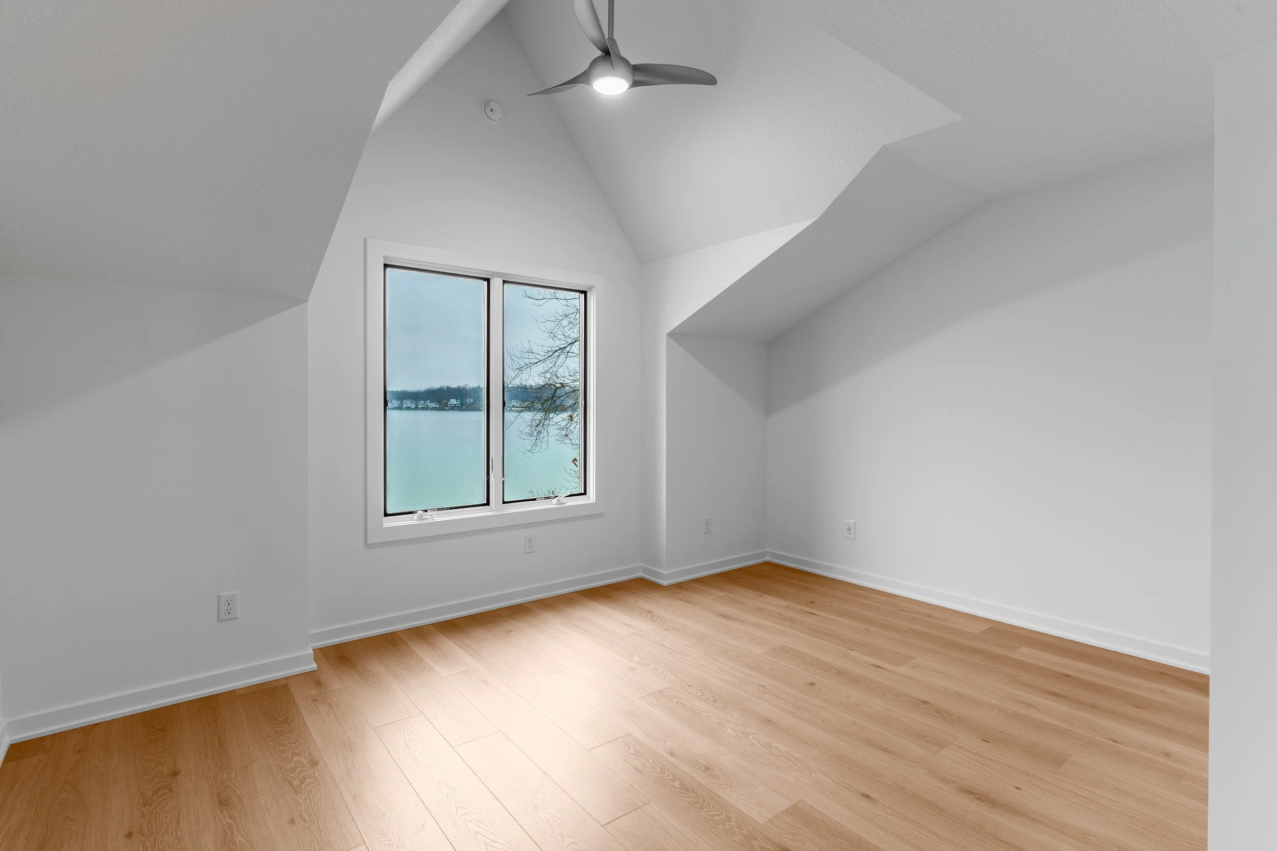 Empty room with a large window overlooking a body of water, white walls, a ceiling fan, and light wood flooring.