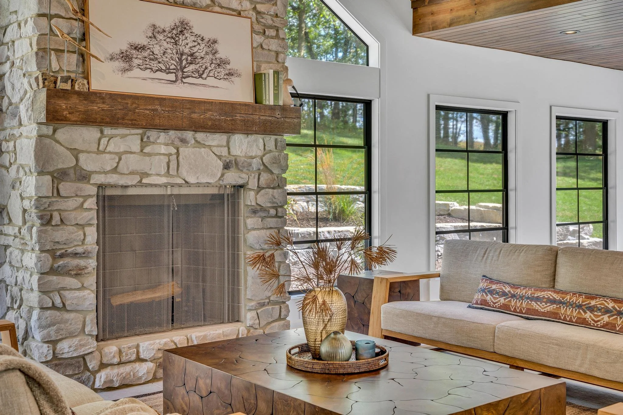Living room with stone fireplace, wooden mantel, art print of a tree, large windows showing outdoor greenery, a beige sofa, and a wooden coffee table with decorative vases and dried plants.