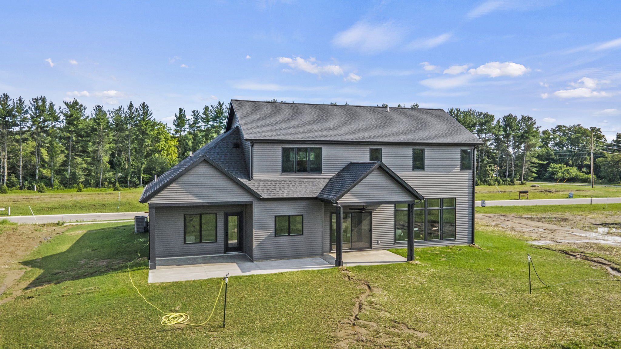 A new two-story house with gray siding and a black roof, situated on a grassy lot with a concrete patio in the backyard, under a blue sky with scattered clouds.
