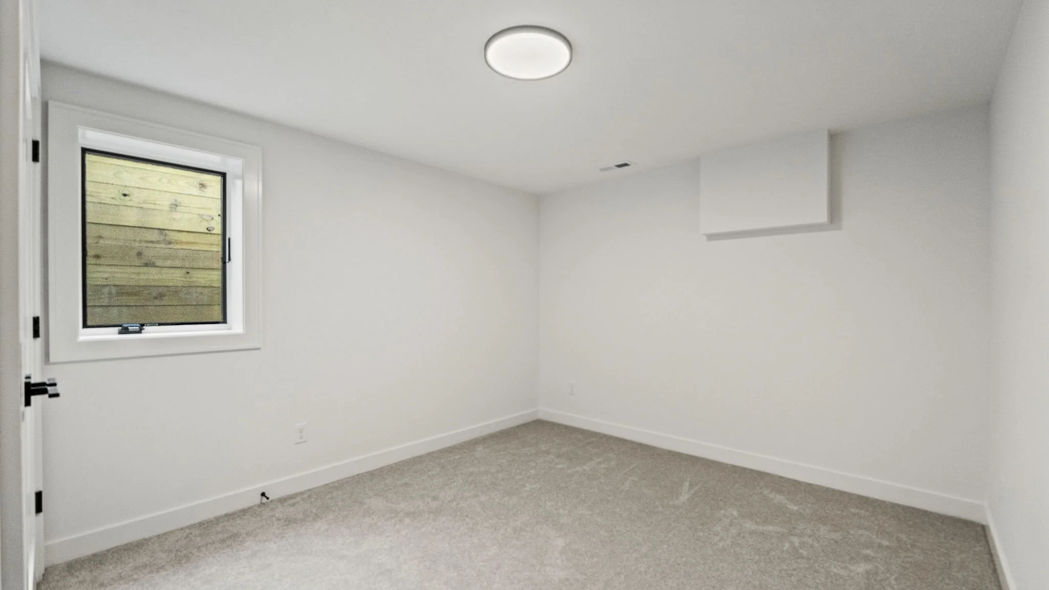 Empty room with beige carpet, white walls, a window with a wooden fence outside, and a ceiling light.