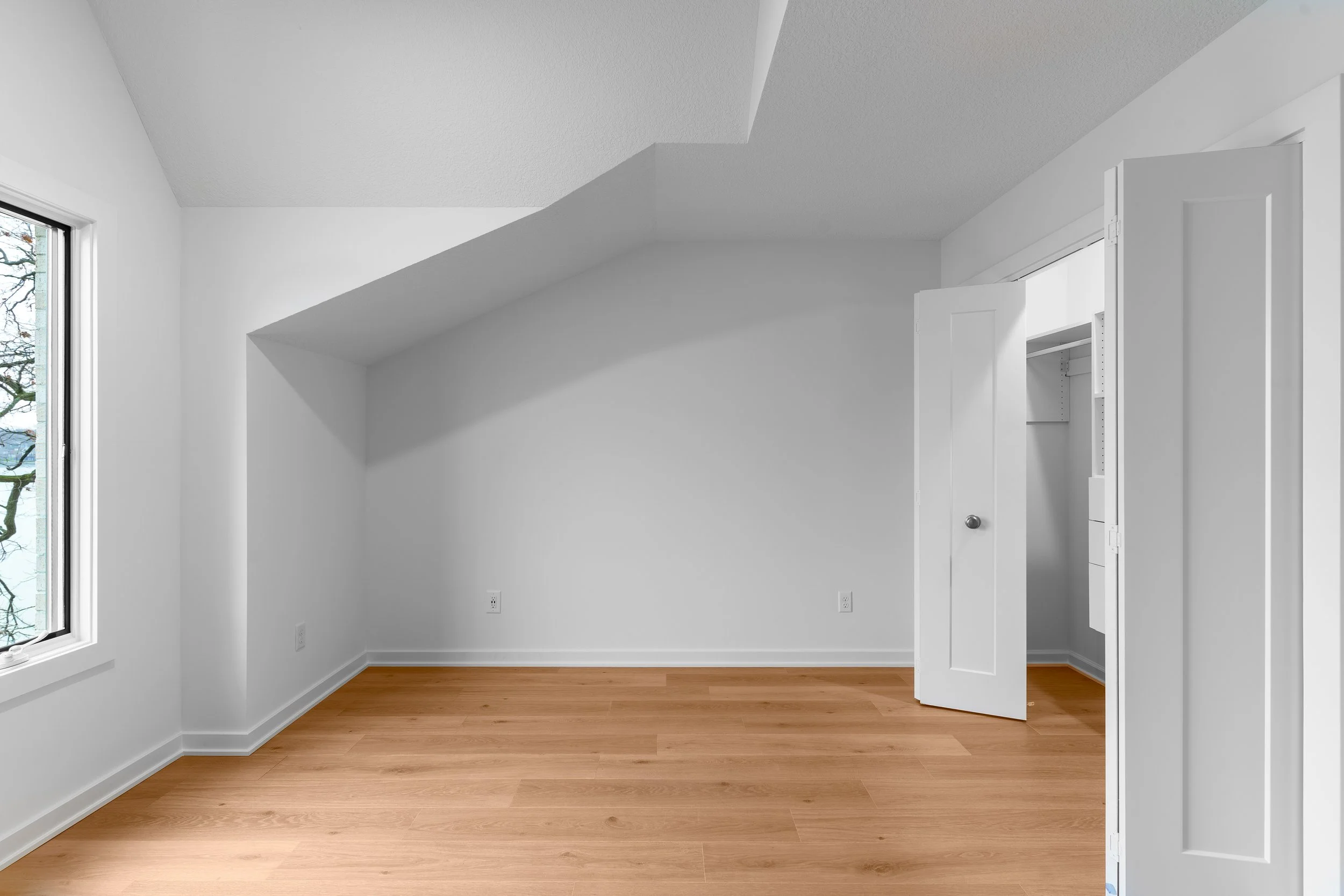 Empty room with white walls, a window showing trees outside, light wooden flooring, and an open closet door.