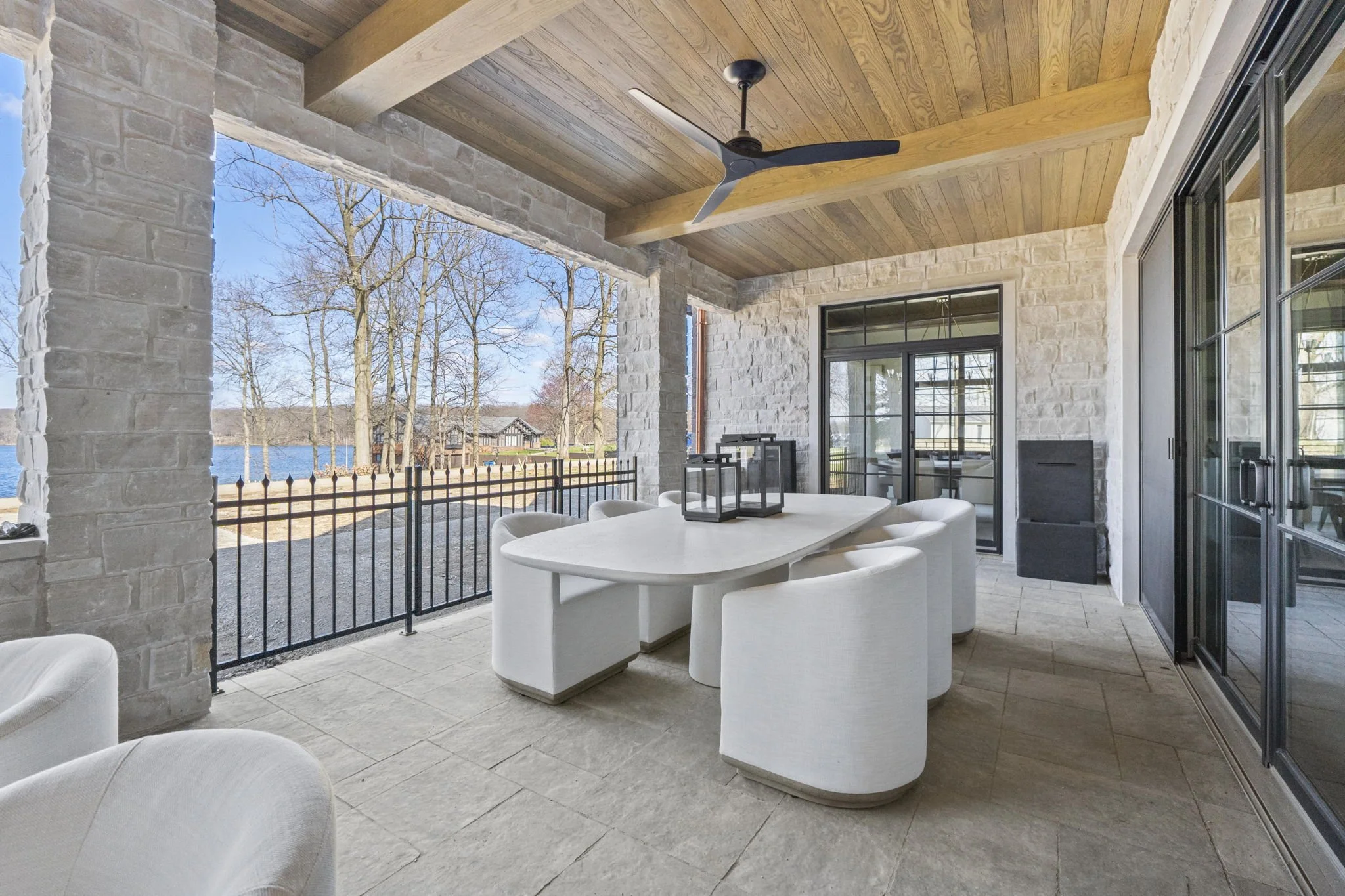 Covered patio with white dining table and chairs, overlooking a lake with trees, stone walls and wooden ceiling, ceiling fan, and glass sliding door.