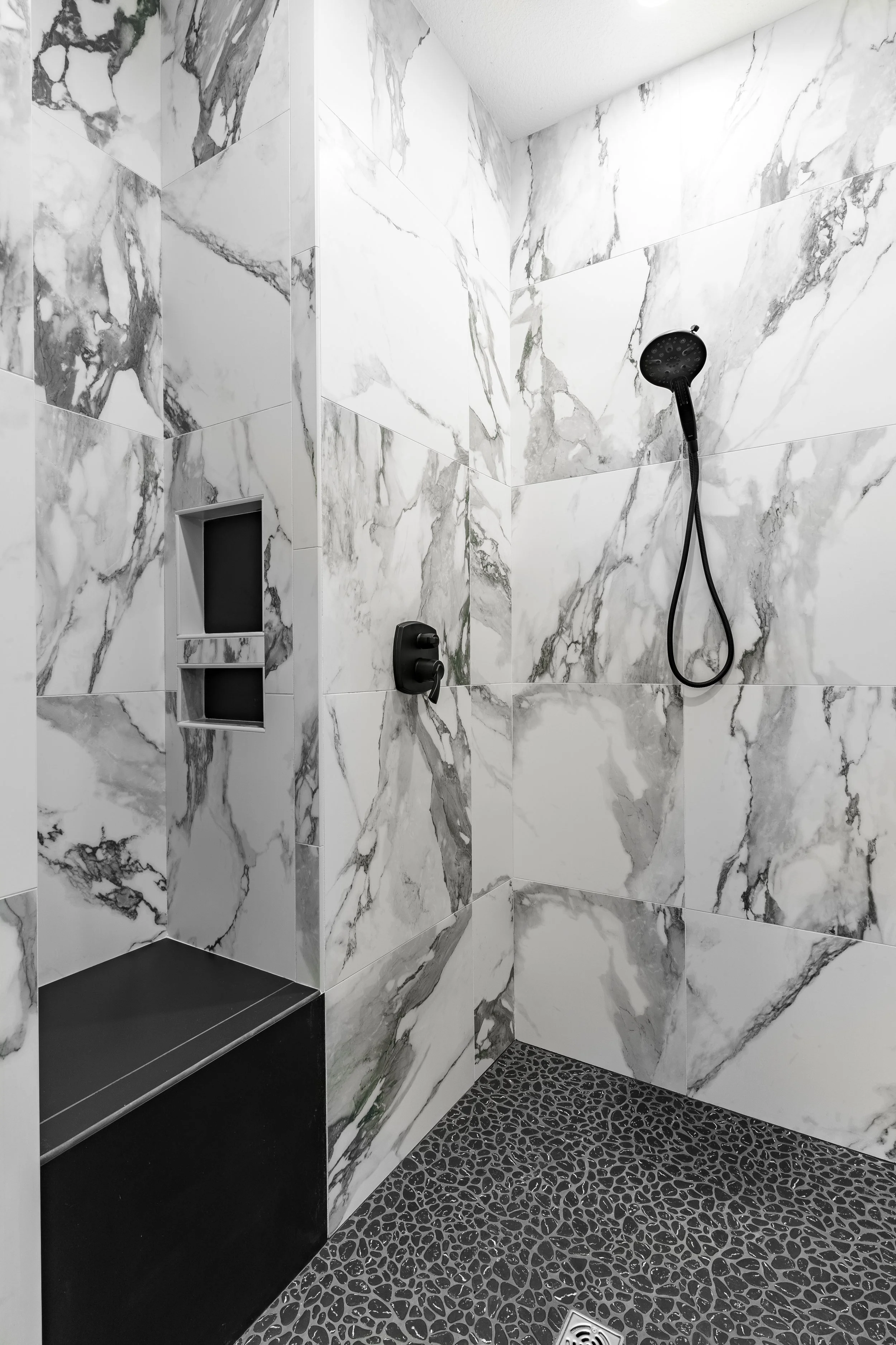 A modern shower with white marble tile walls, a black shower head, a handheld shower, a built-in soap niche, and a pebble tile floor.