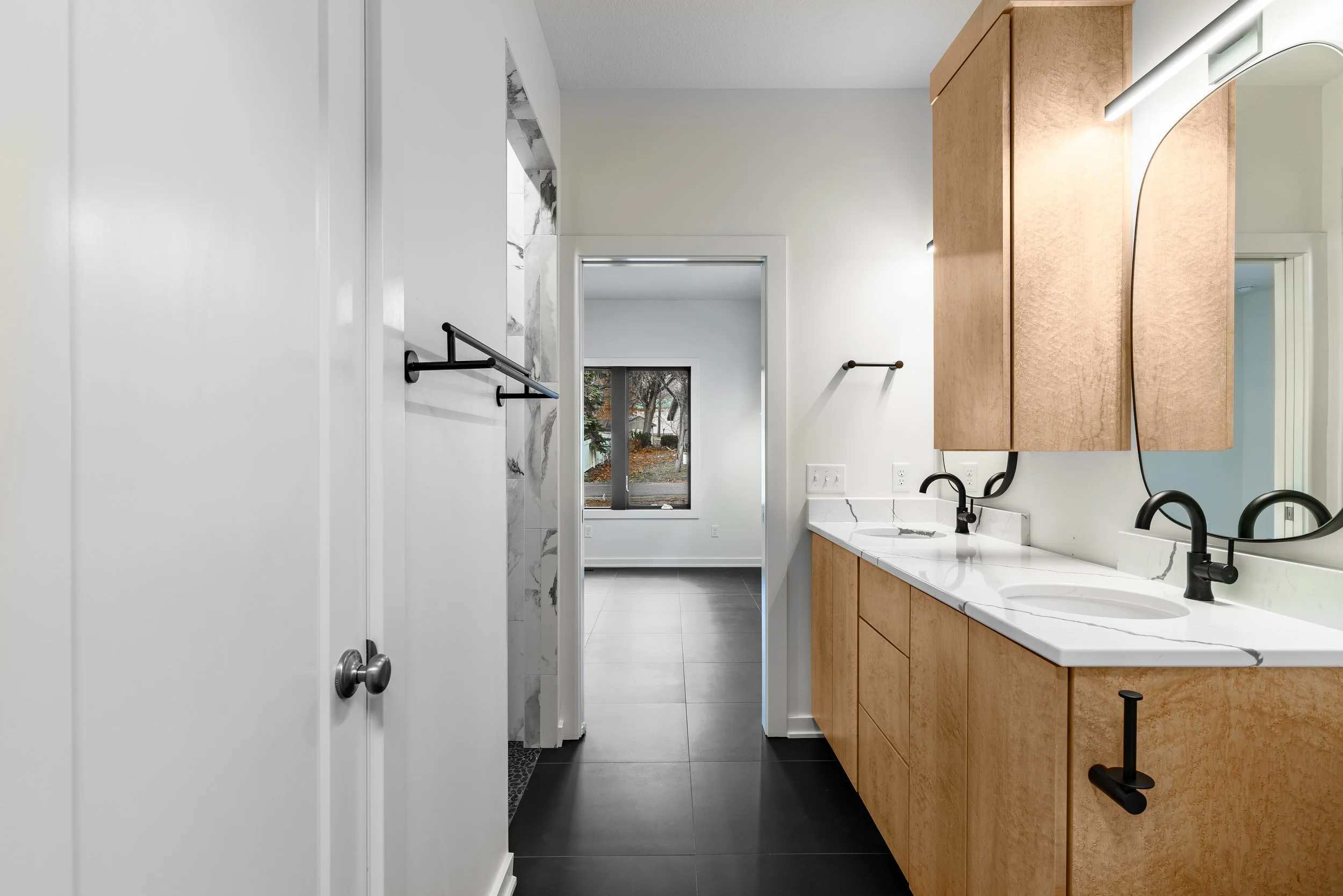 Bathroom with dual sinks, black faucets, wood cabinets, and large mirror, with a view into another room with window.