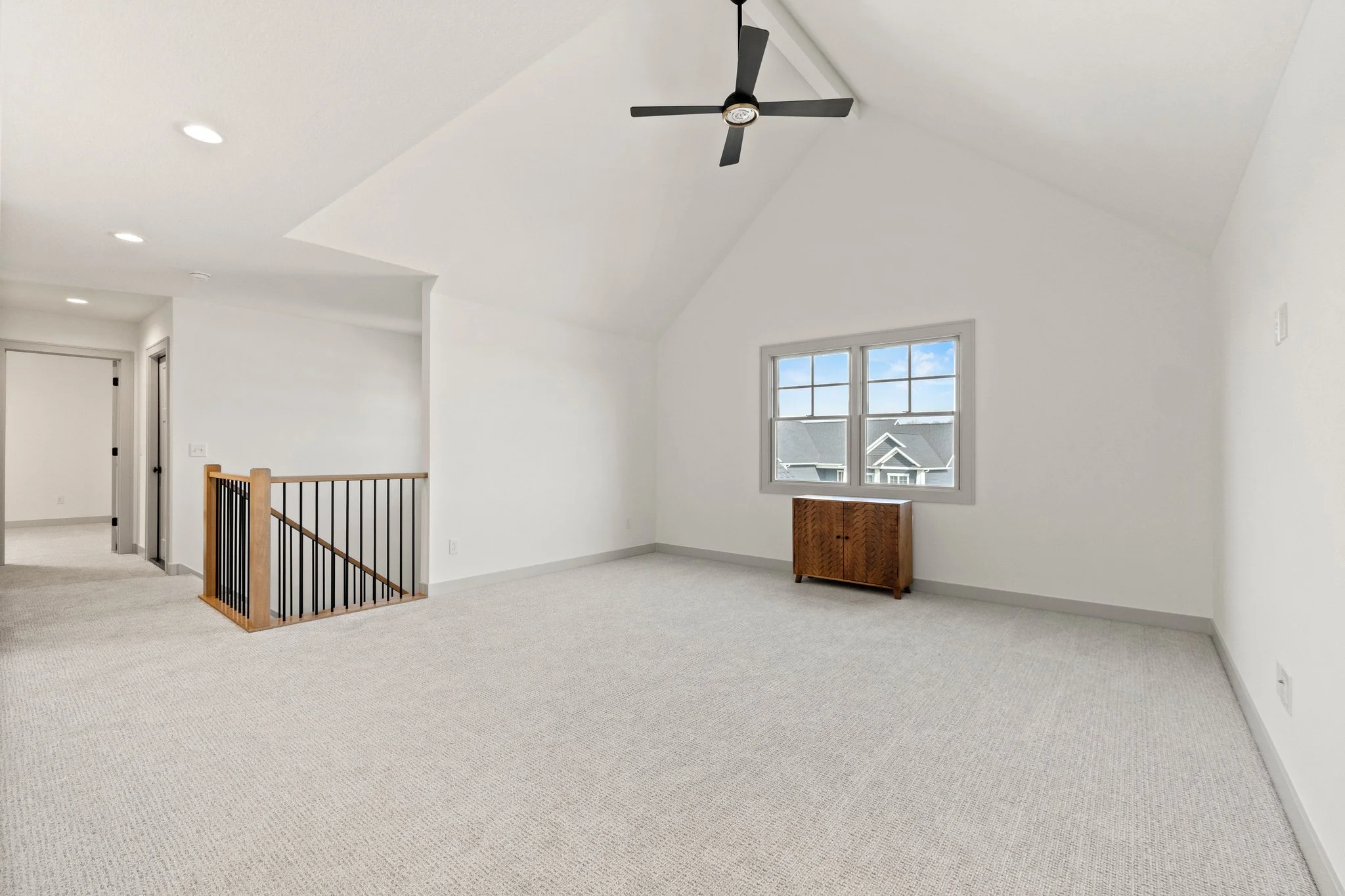 Empty attic room with white walls, beige carpet, a ceiling fan, and a window showing neighboring rooftops.