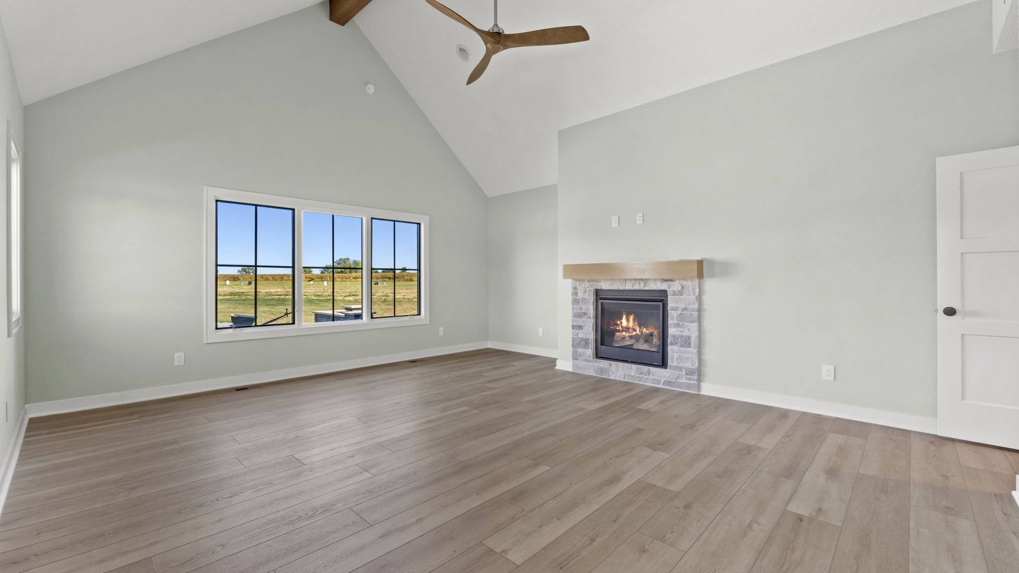 Empty living room with large window, white walls, wood flooring, a fireplace with a stone surround, and a ceiling fan.
