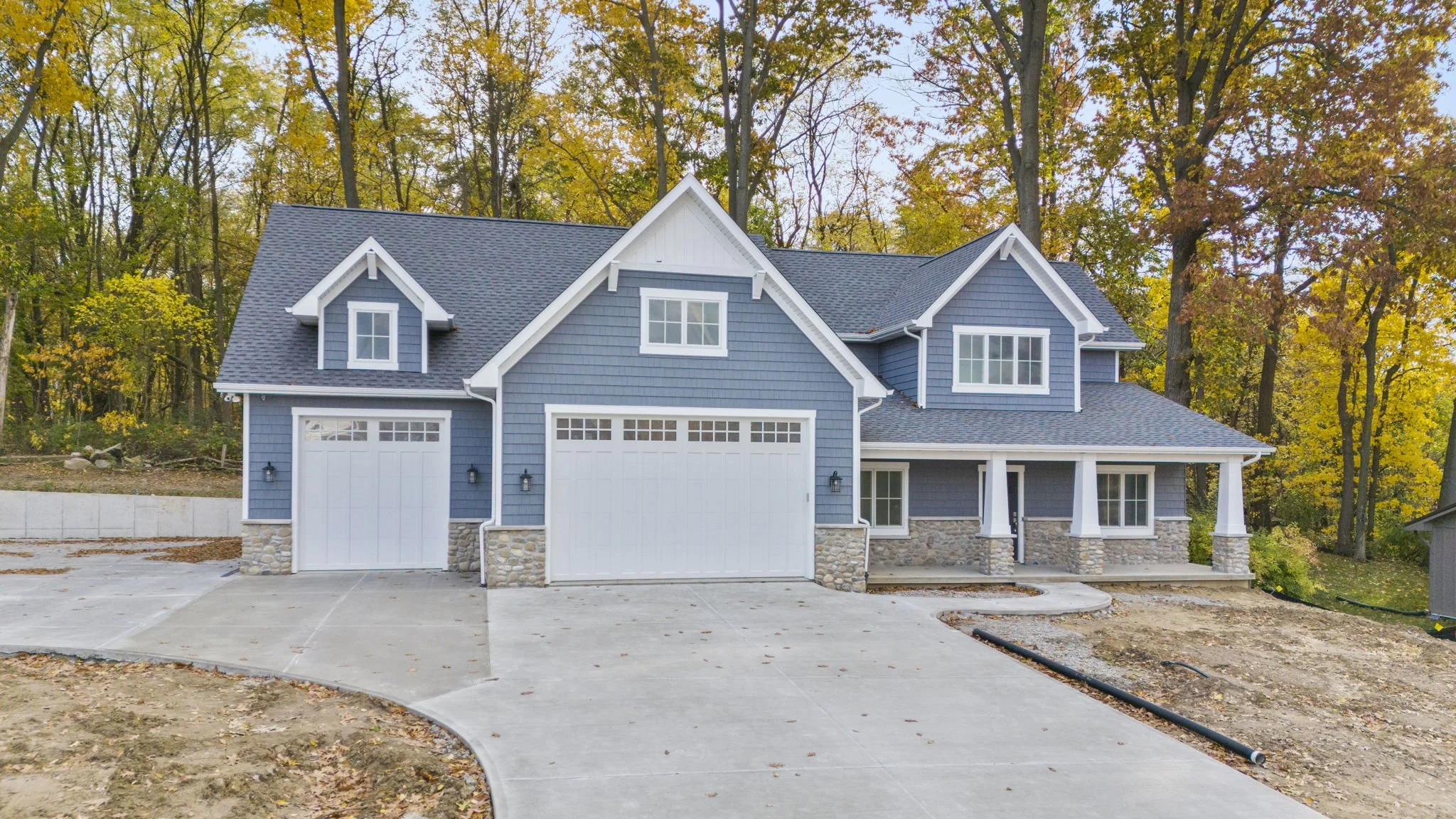 New blue two-story house with a front porch, two garage doors, surrounded by trees with fall foliage.