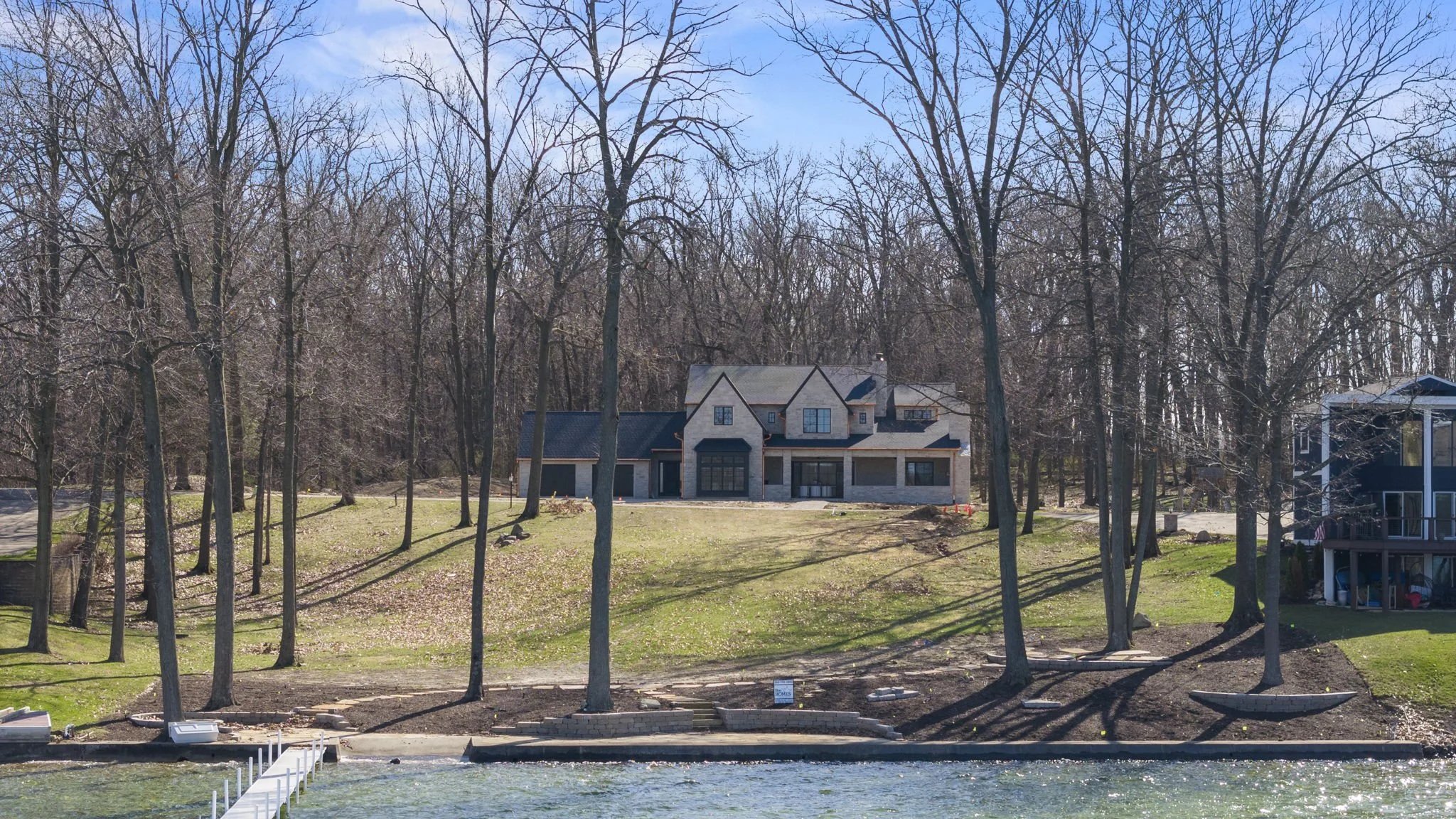 A modern house on a grassy hill by a body of water, with leafless trees and a partly cloudy sky.