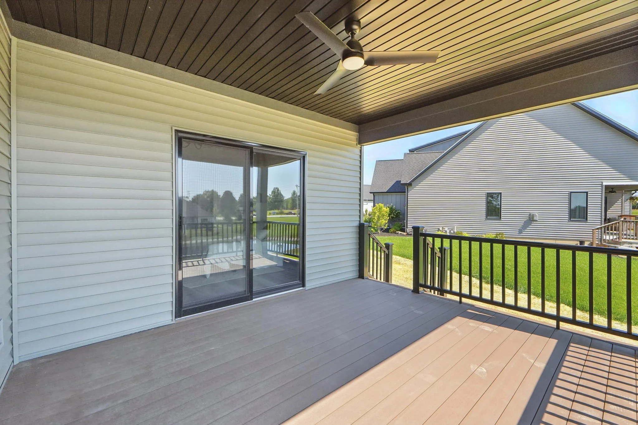 Empty outdoor balcony with wooden flooring, a ceiling fan, and a sliding glass door, overlooking neighboring houses and green lawns.