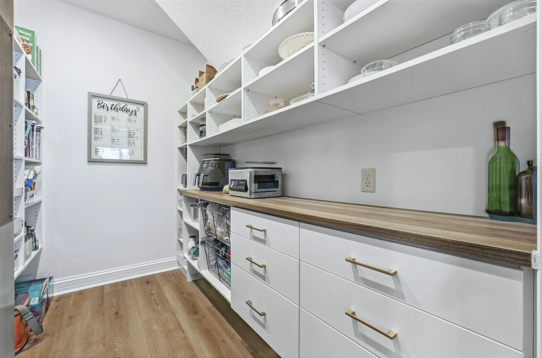 A pantry with white shelves filled with plates, bowls, and glassware. A wooden countertop with small appliances like a toaster and microwave. Drawers with gold handles, a wall socket, and a framed birthday calendar on the wall.