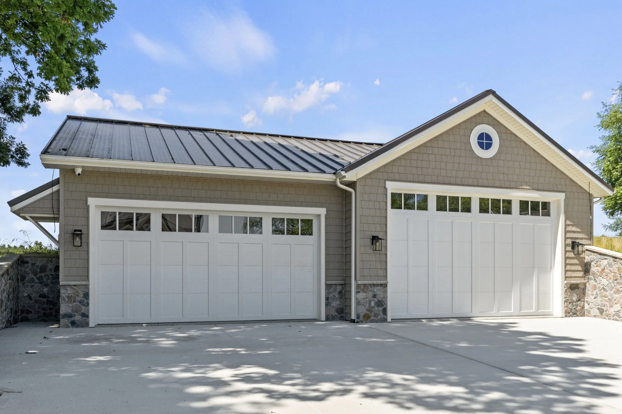 Two-car garage with white doors and a stone foundation, beige siding, gabled roof, and black side lights, under a blue sky with scattered clouds.