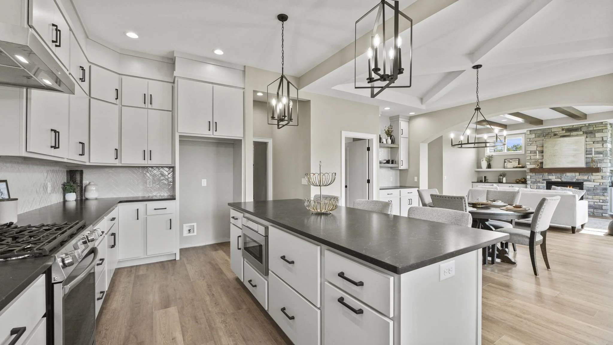 Modern open-plan kitchen and living room with white cabinets, black countertops, wooden flooring, and a stone fireplace.