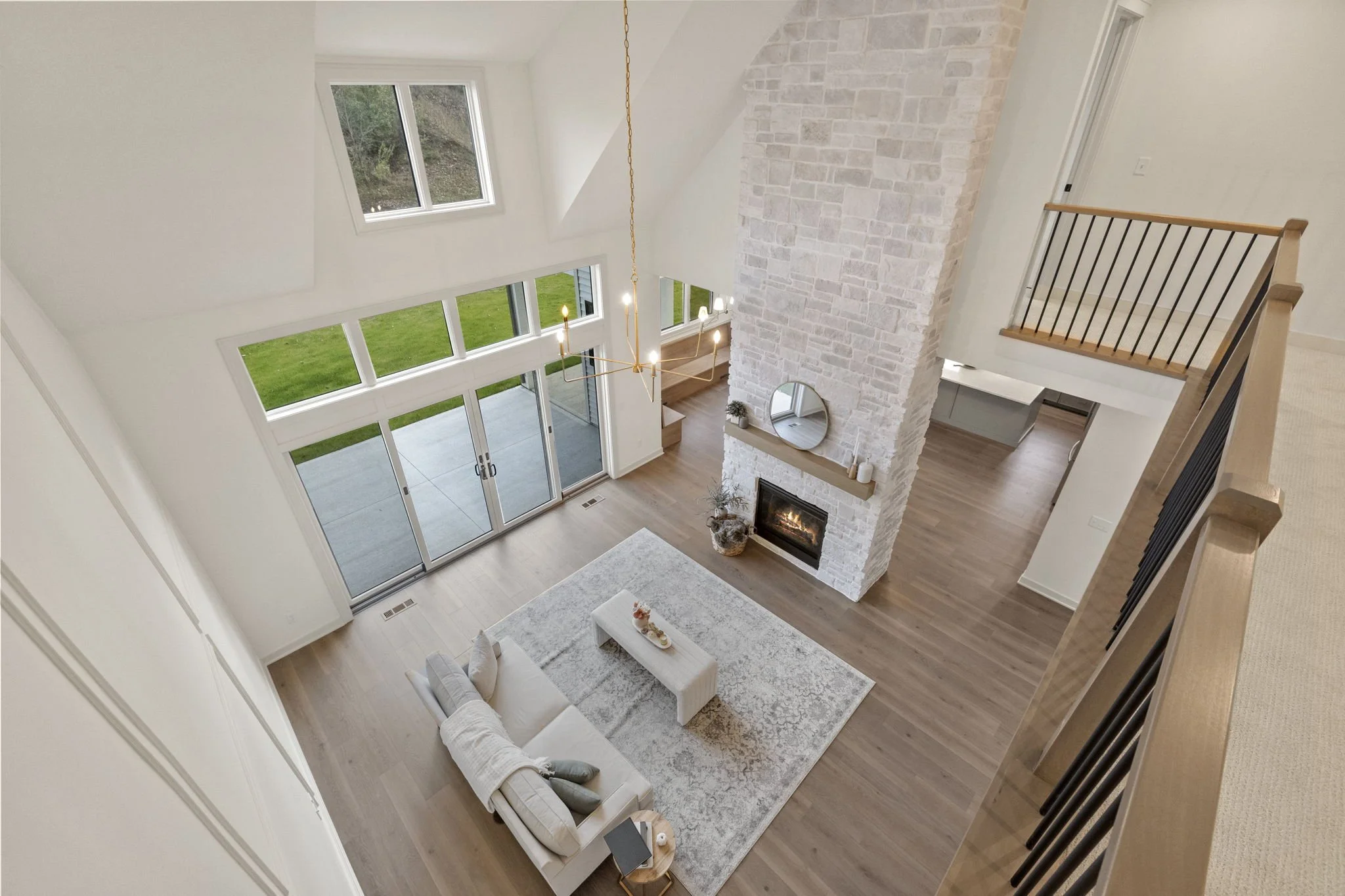View of a modern living room from an upper floor, showing a large white sofa with pillows, a light-colored area rug, a fireplace with a white brick chimney, a round mirror above the fireplace, a minimalist chandelier hanging from the ceiling, and lar