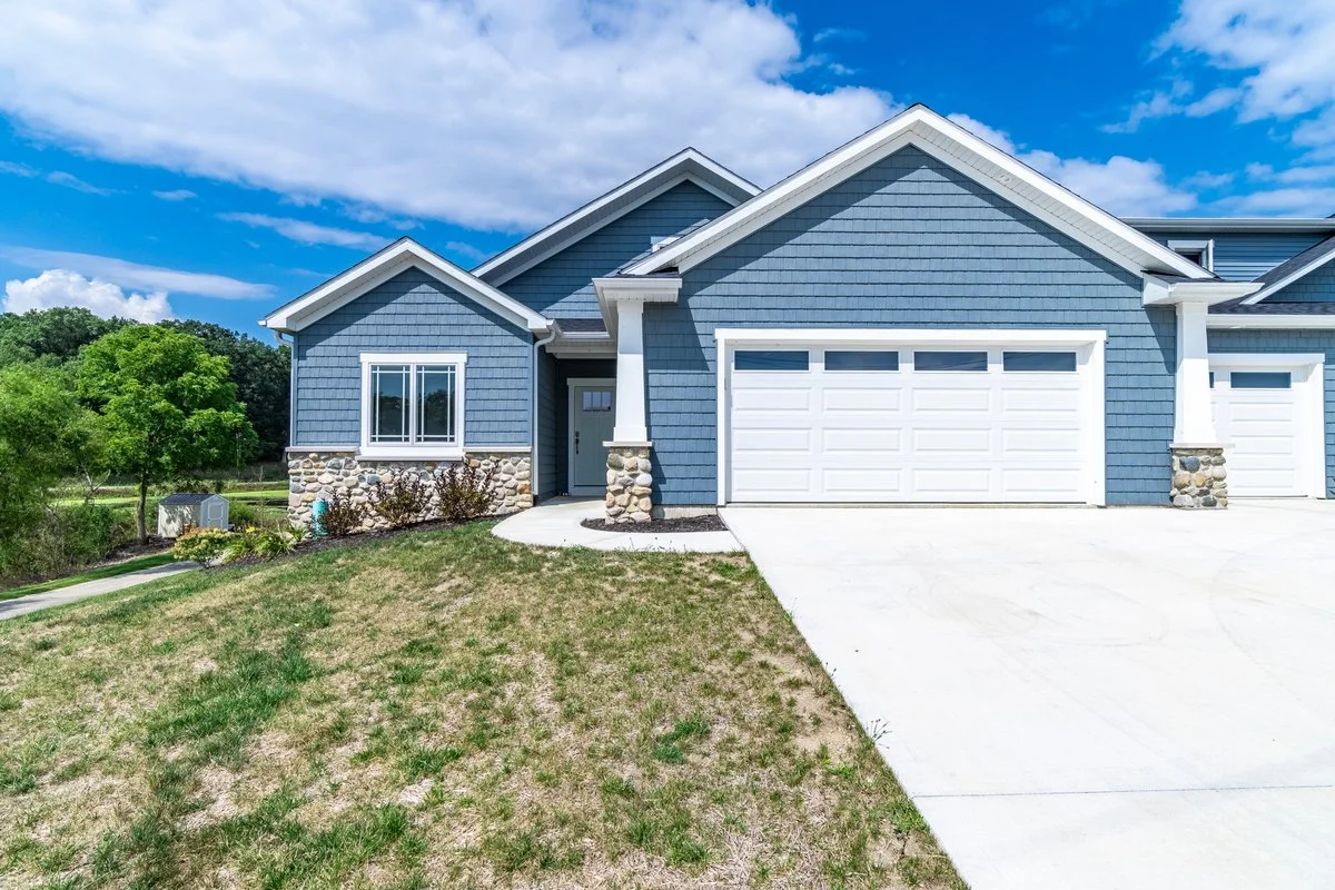 A blue single-family house with a white garage door, stone accents at the base of the house, and a small front porch area, set against a cloudy sky with green trees in the background.