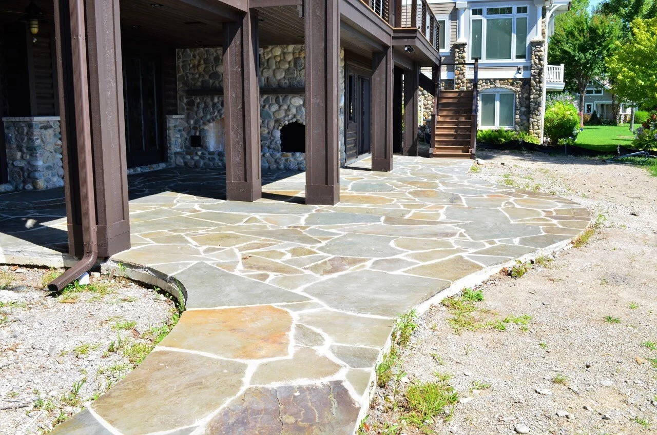 A newly paved stone patio in front of a house with a wood and stone exterior, surrounded by some grass and dirt.
