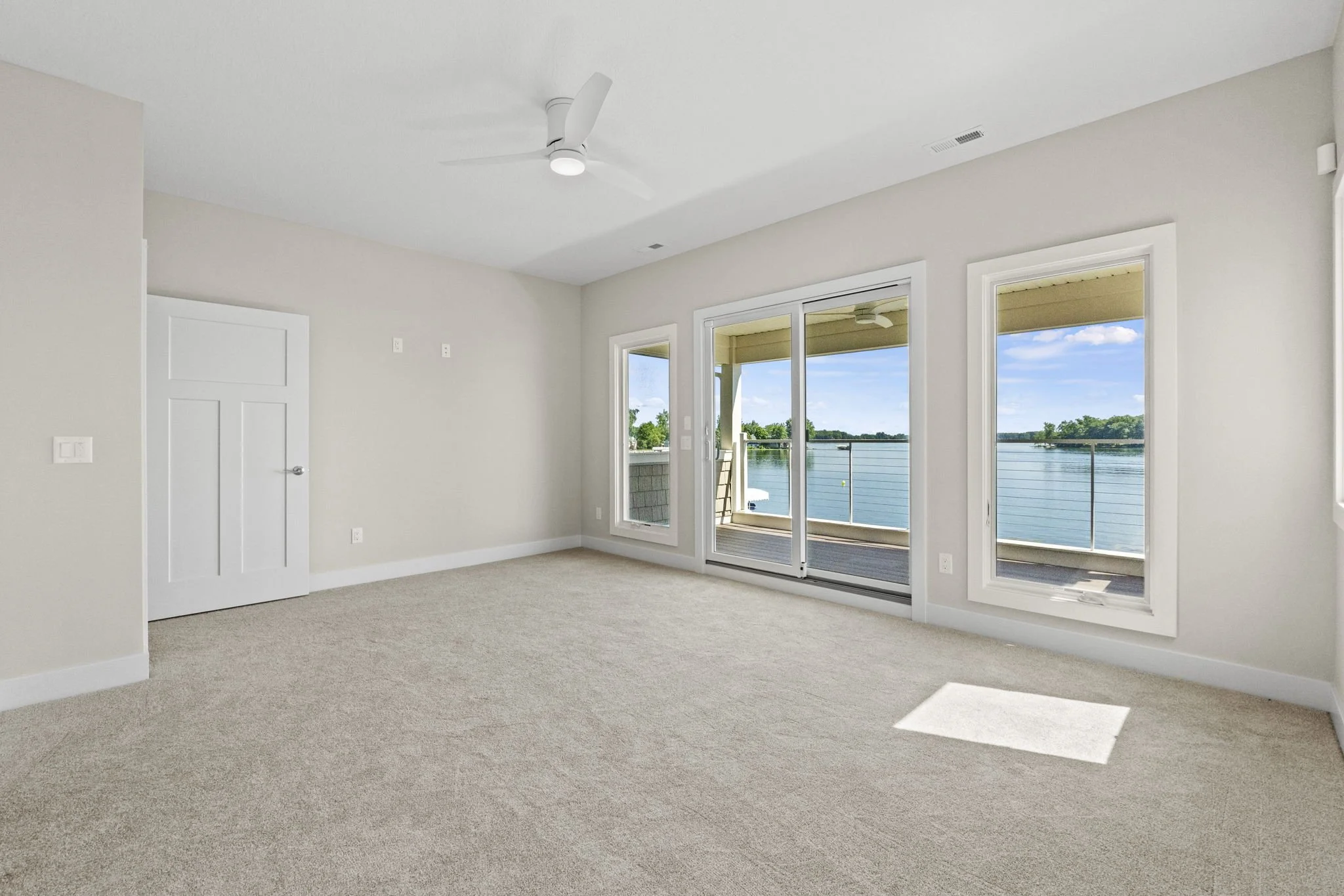 Empty living room with beige carpet, white walls, large windows, and a sliding door leading to a balcony with water view.