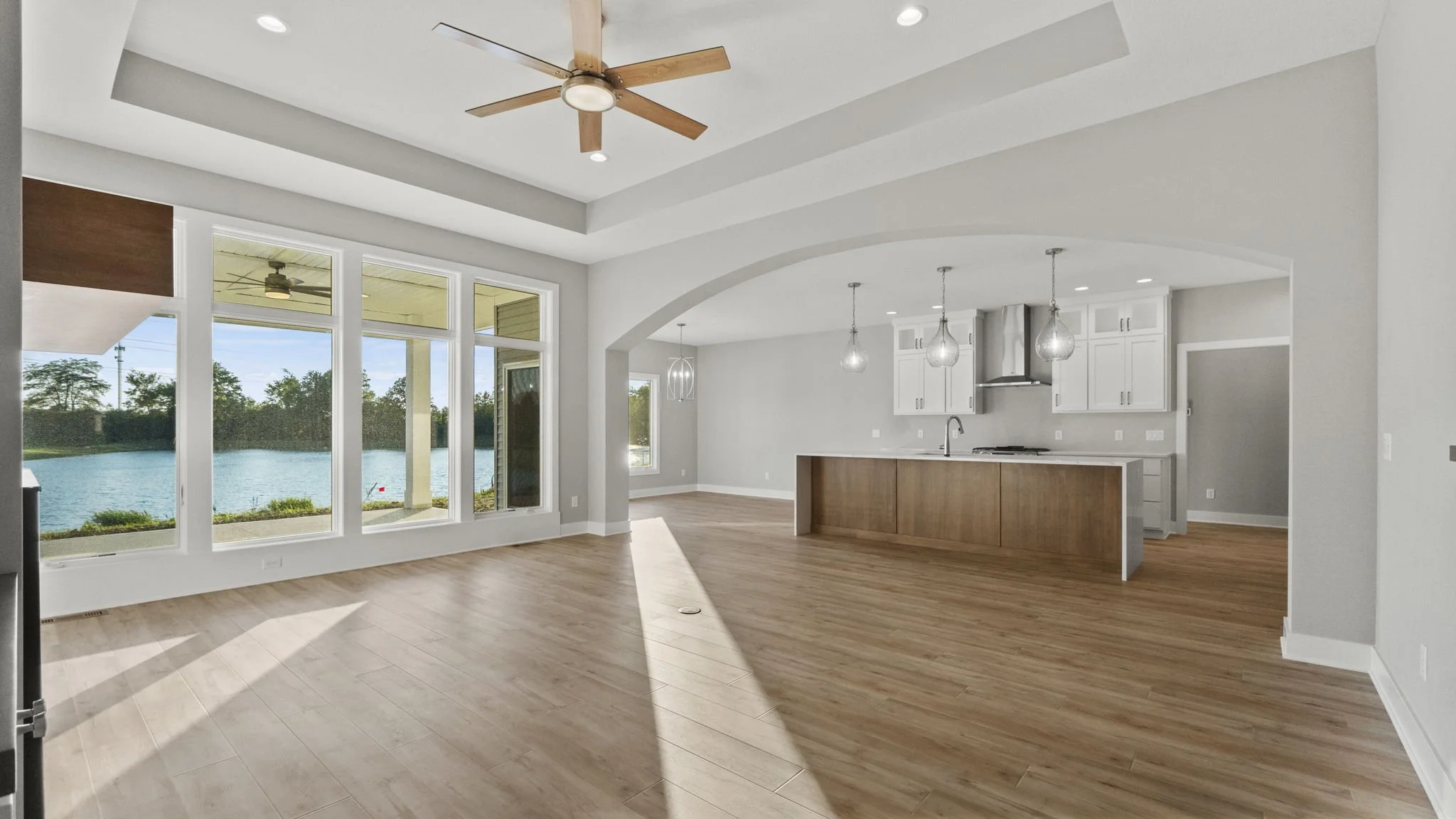 Interior view of a modern open-concept living room and kitchen with large windows overlooking a lake, white walls, wood flooring, a ceiling fan, pendant lights over the kitchen island, and white cabinets with stainless steel appliances.