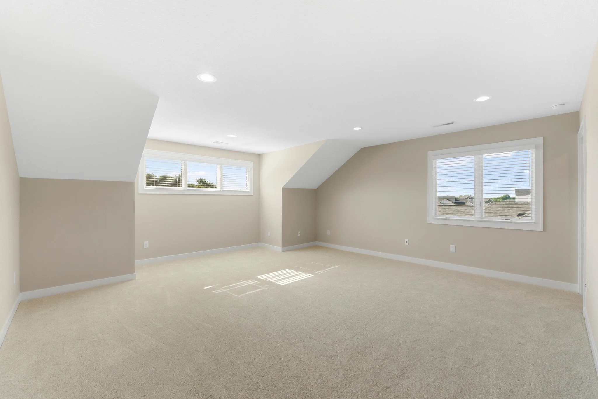 Empty attic room with beige walls, white ceiling with recessed lights, two large windows with blinds, and beige carpet flooring.