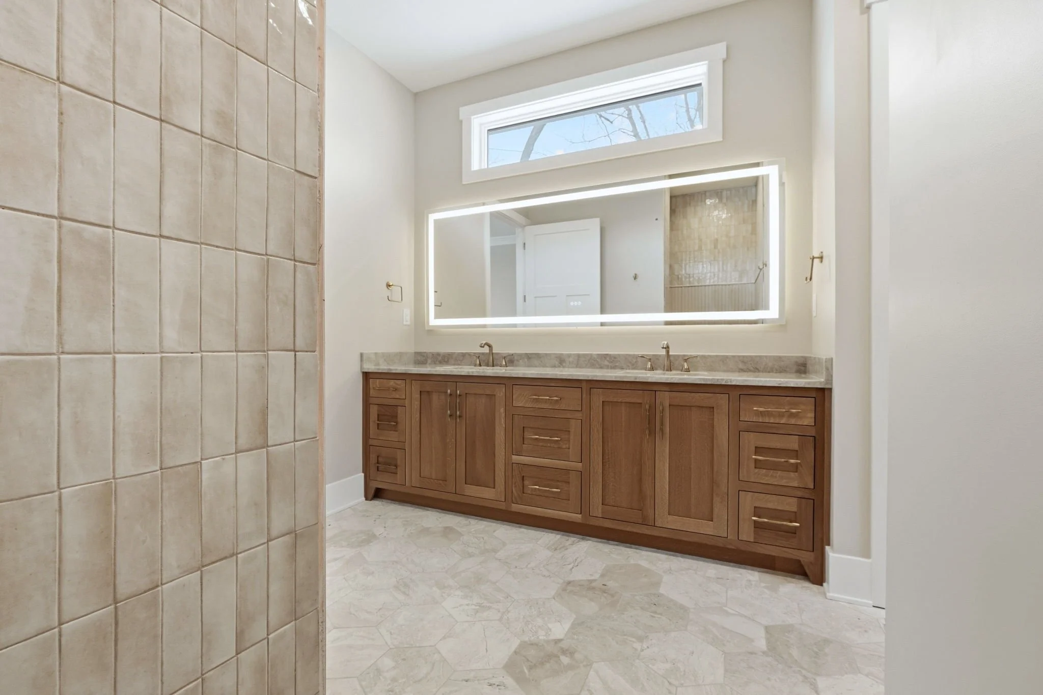 Bathroom with a large illuminated mirror above a wooden vanity with two sinks, a granite countertop, beige tiled floor, and a window above the mirror.