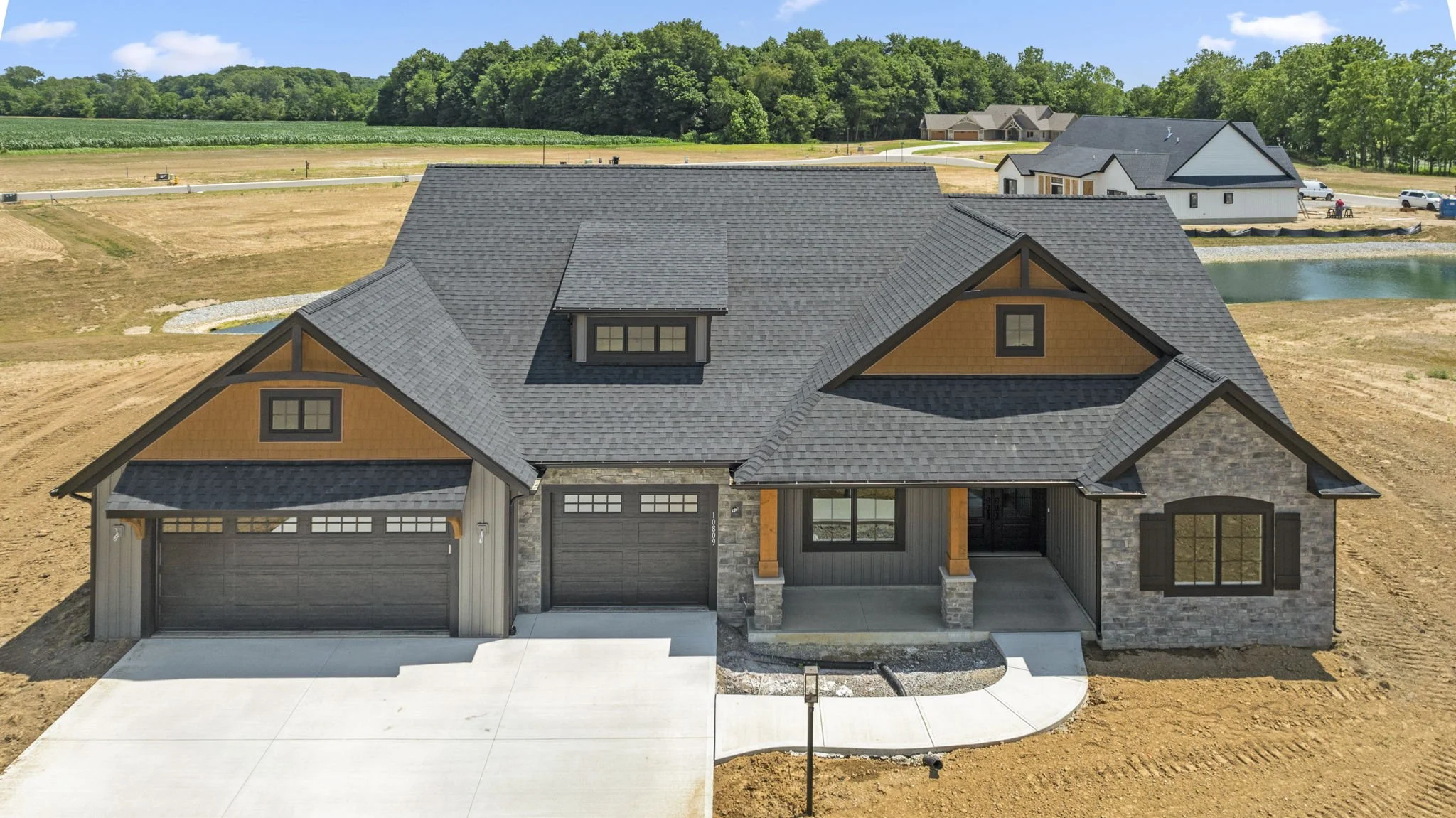 Modern house under construction with gray roof, stone and wooden accents, two-car garage, and front porch, on a cleared lot with construction equipment and neighboring houses in the background.