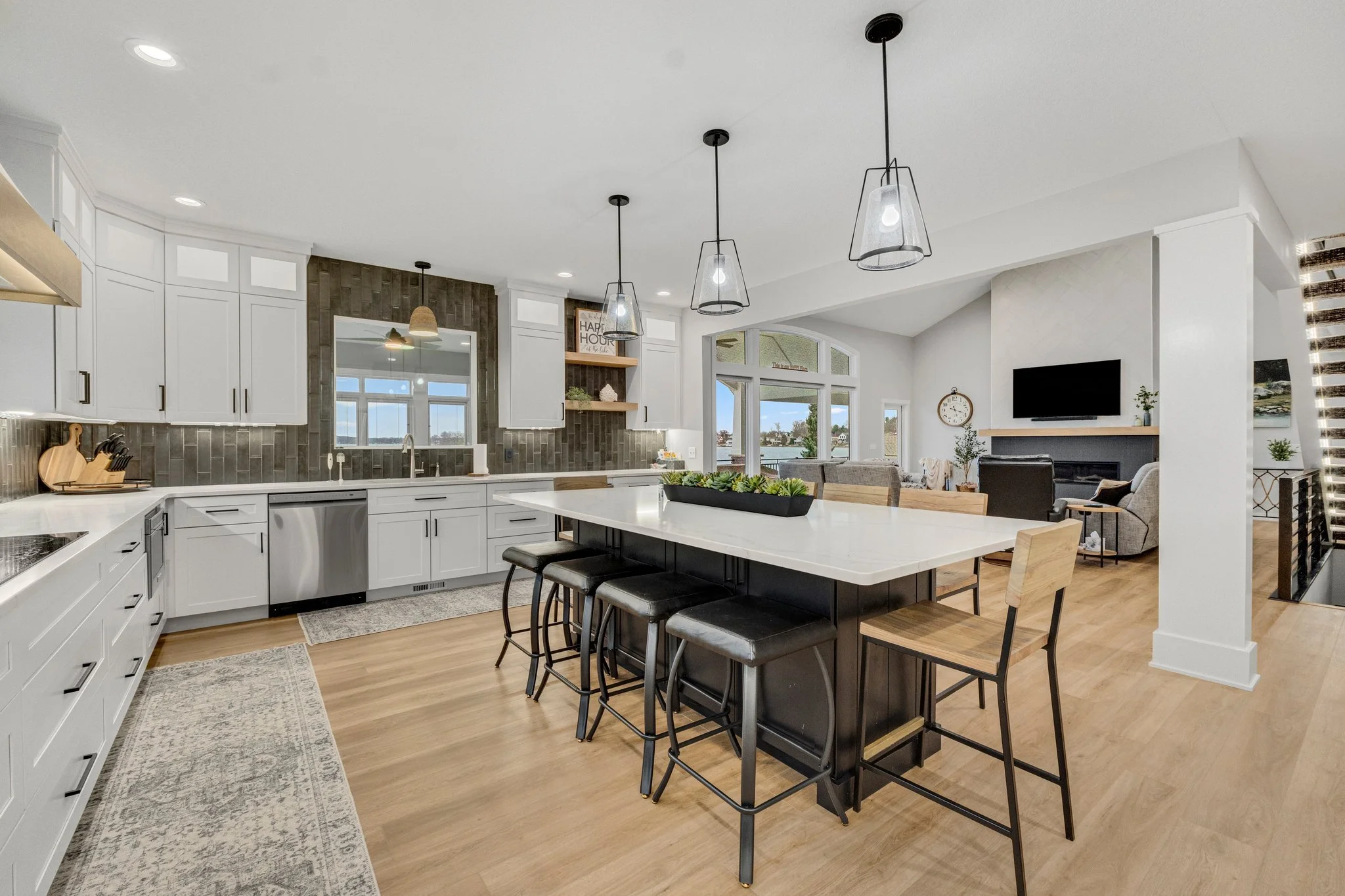 Open-concept kitchen and living room with white cabinets, gray backsplash, large kitchen island with black base and white countertop, pendant lighting, and a seating area with television and fireplace.