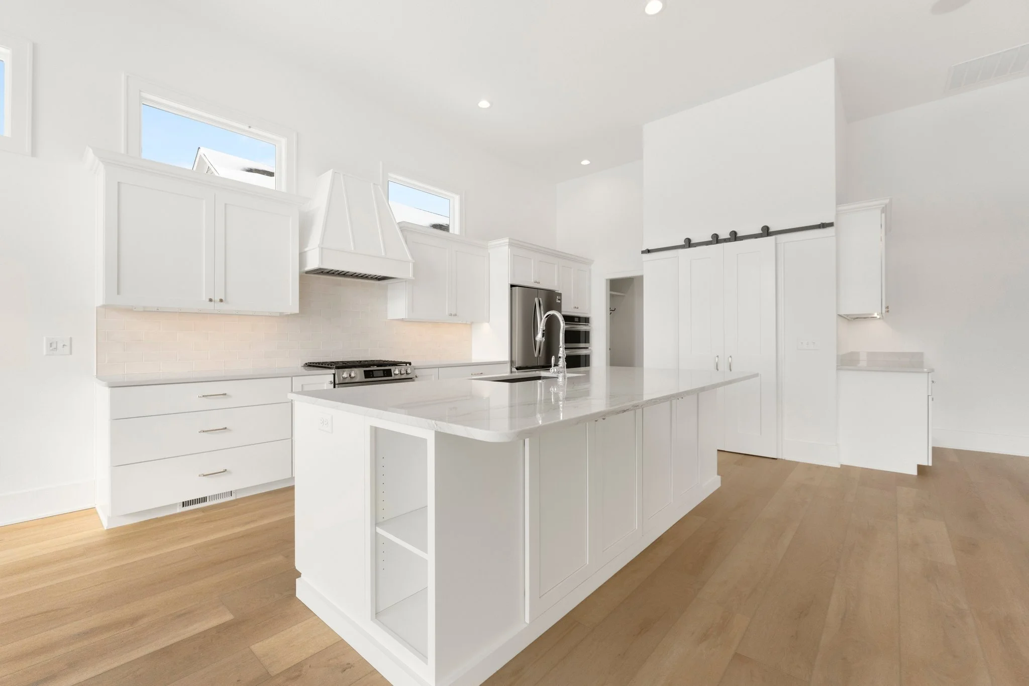 Empty modern kitchen with white cabinets, a central island with a built-in sink, stainless steel appliances, wood flooring, and three small windows above the cabinets.
