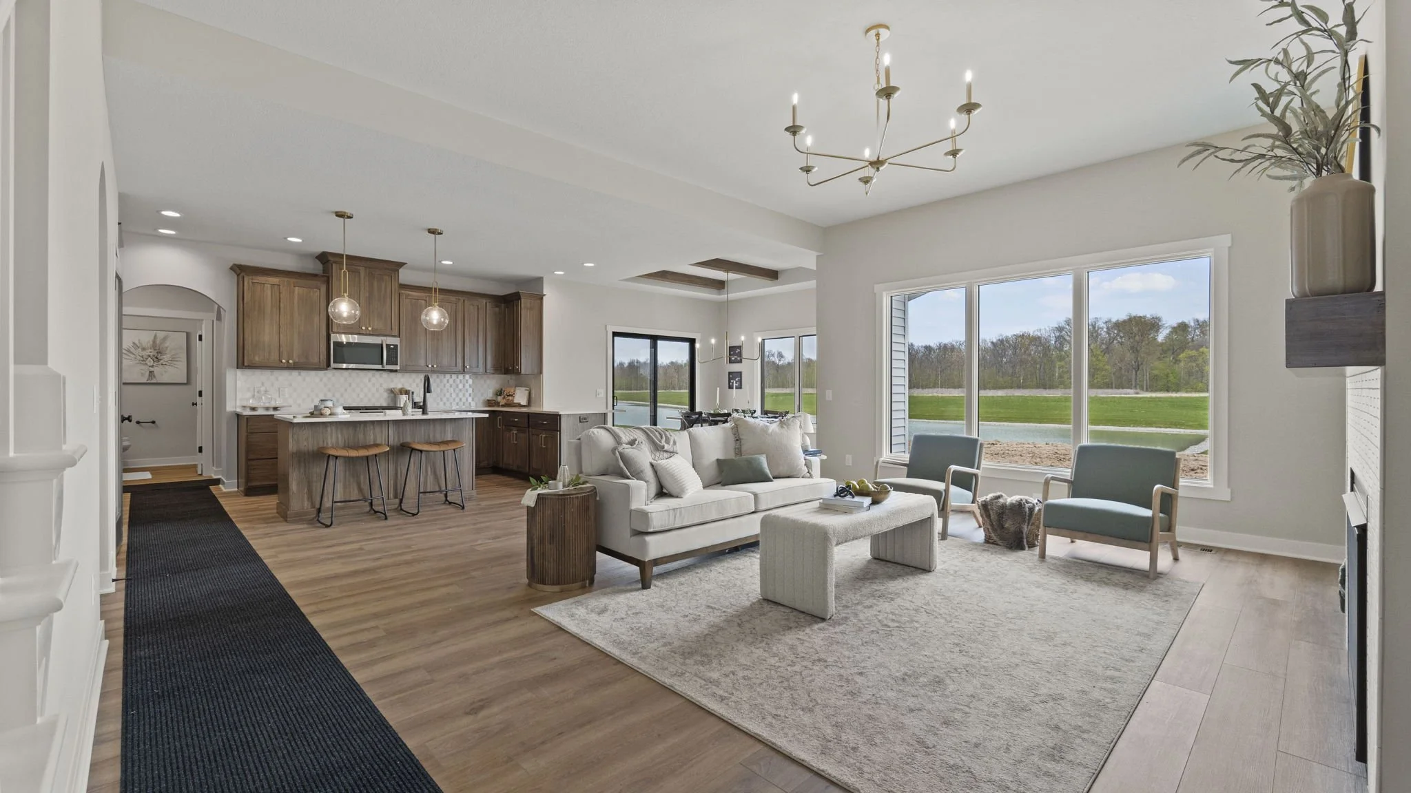 Open concept living area with large windows showing green outdoor scenery, featuring a white sofa, two blue armchairs, a wooden kitchen island, and modern chandelier lighting.