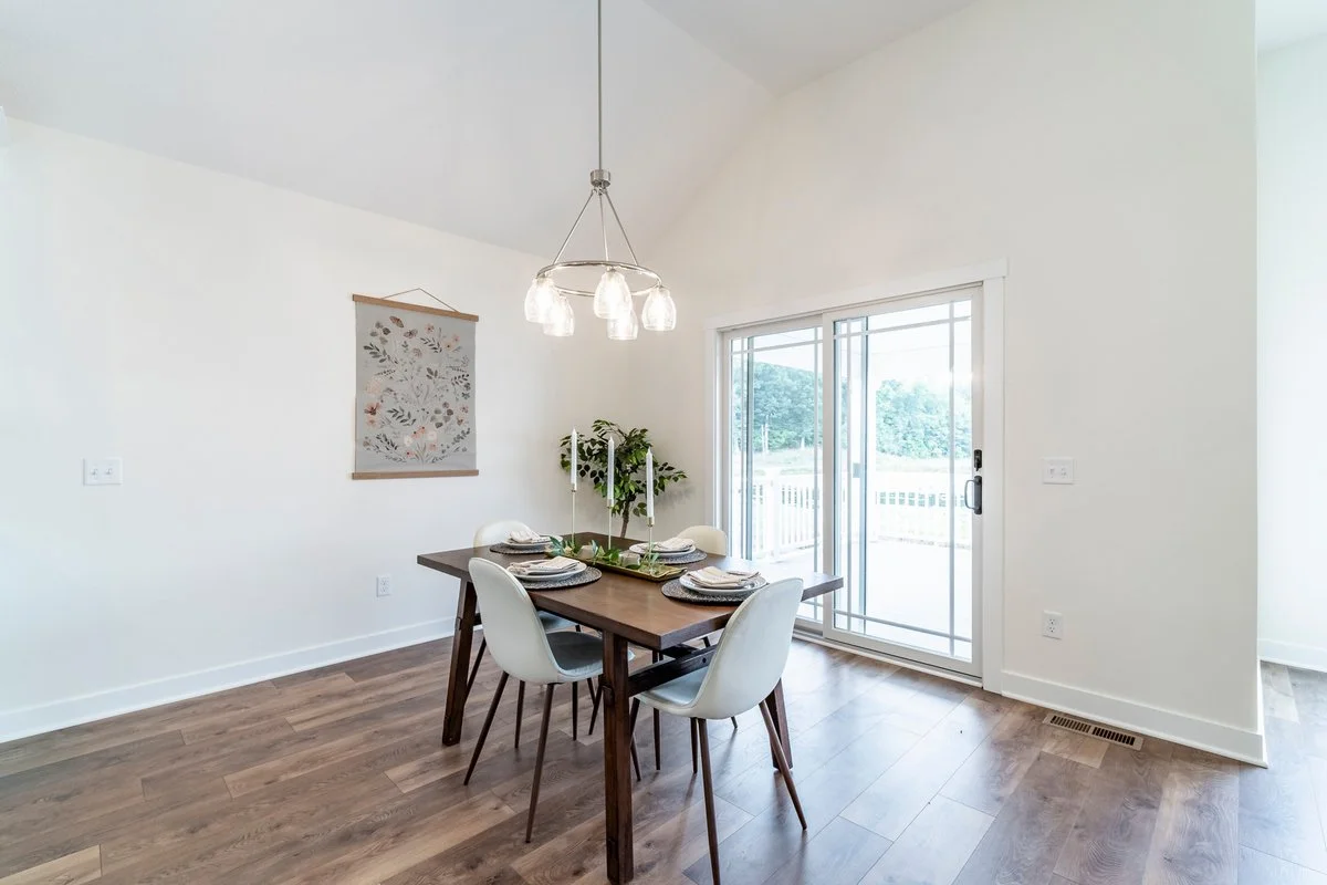 A dining area with a wooden table set for four, white chairs, a pendant light hanging above, a sliding glass door to the outside, a potted plant, and wall decor in a bright room with wooden flooring.