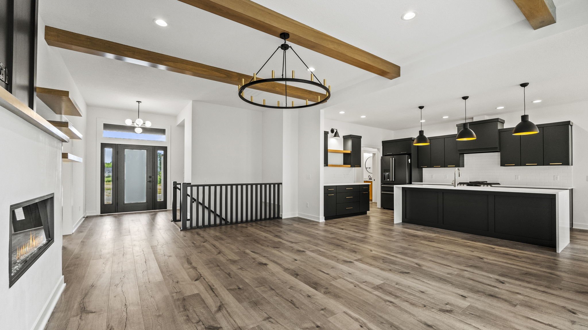 Interior of a modern open-concept kitchen and living space with black cabinets, a central island, wooden beams, pendant lighting, and a front door with windows.