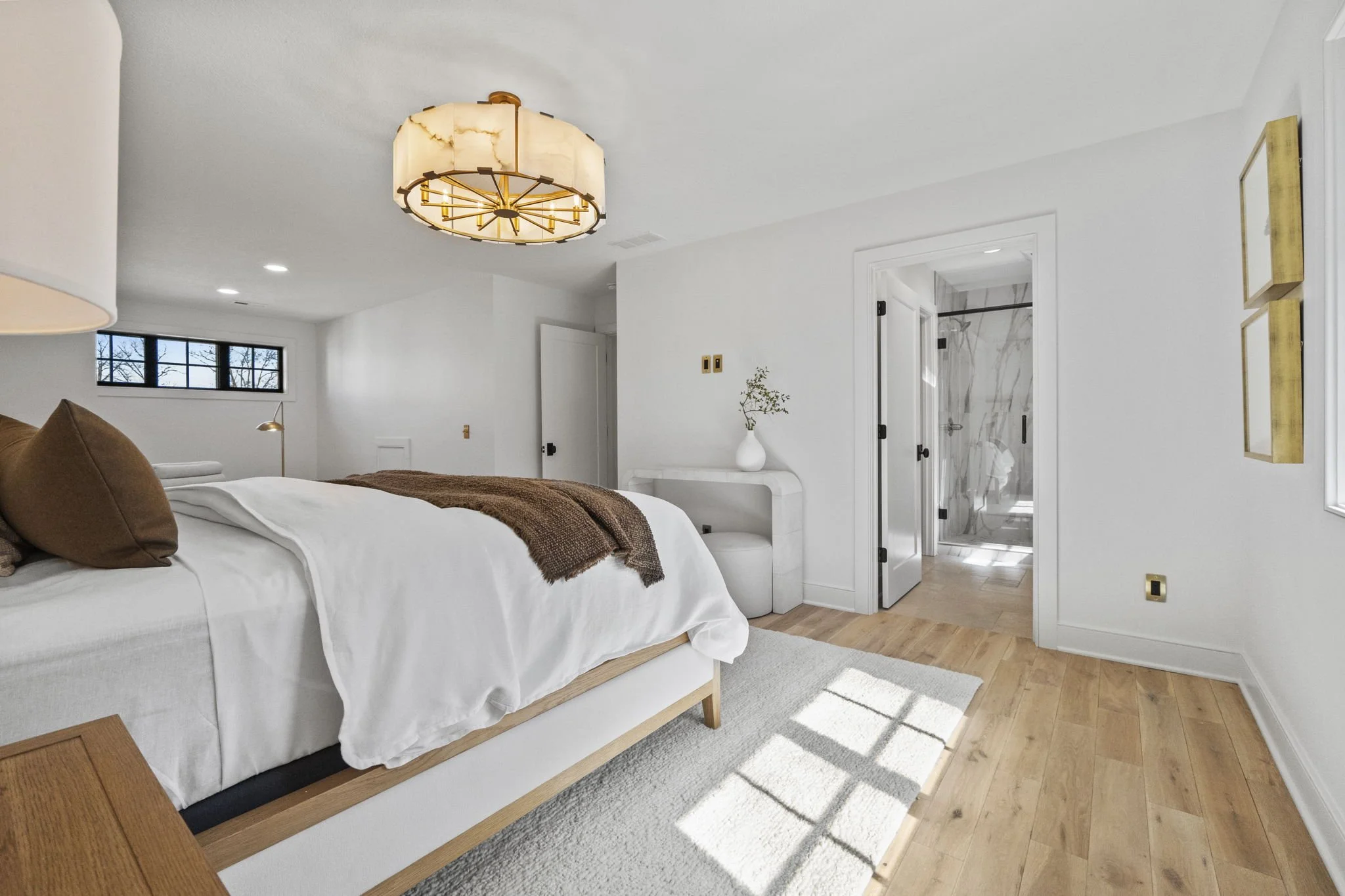 A modern bedroom with white walls, wooden flooring, and a white bed with pillows and a brown blanket. There is a window with black frames, a white vase with greenery on a white curved table, and two gold wall art pieces. The room leads to a bathroom 