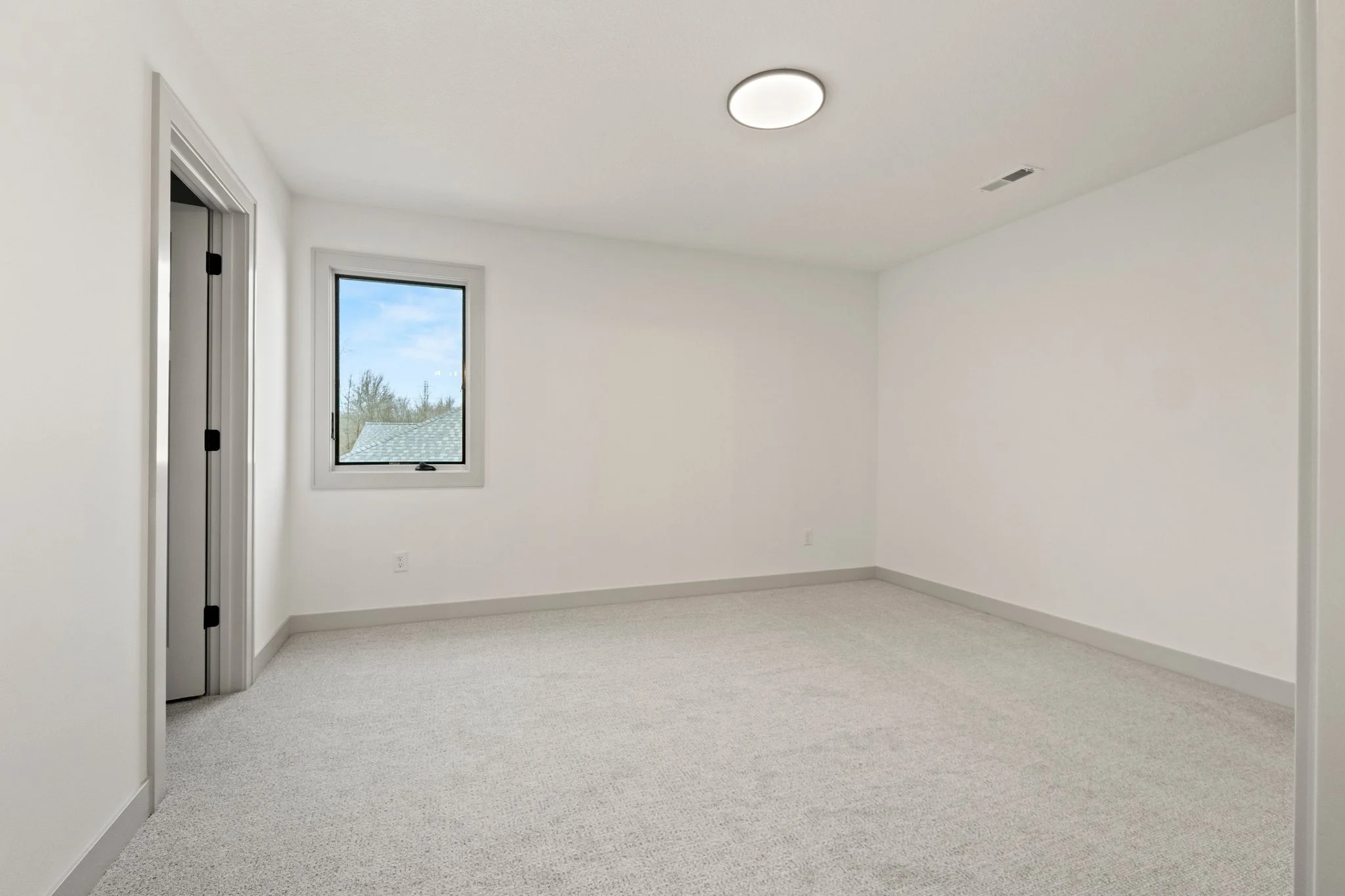 Empty room with white walls, a window showing a view of the sky and trees, and a ceiling light.