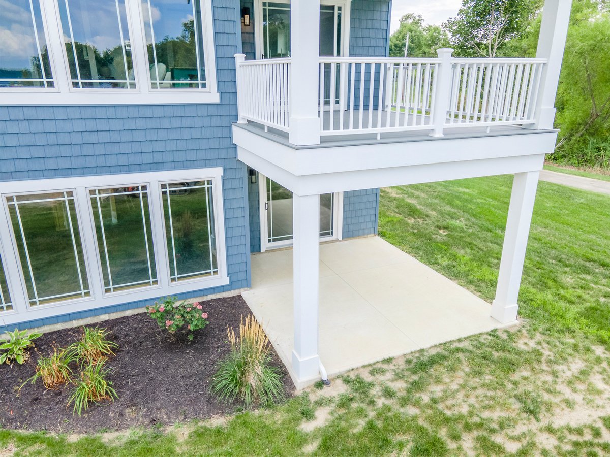 Exterior view of a blue house with a second-story balcony with white railing and a ground-level concrete patio, surrounded by a lawn and garden bed with plants and flowers.