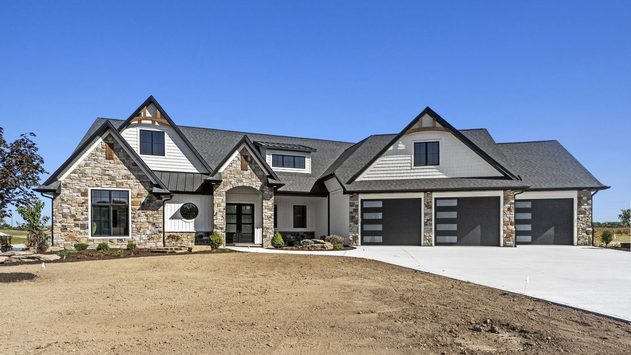 Newly constructed house with stone and white siding, black garage doors, and a concrete driveway under a clear blue sky.