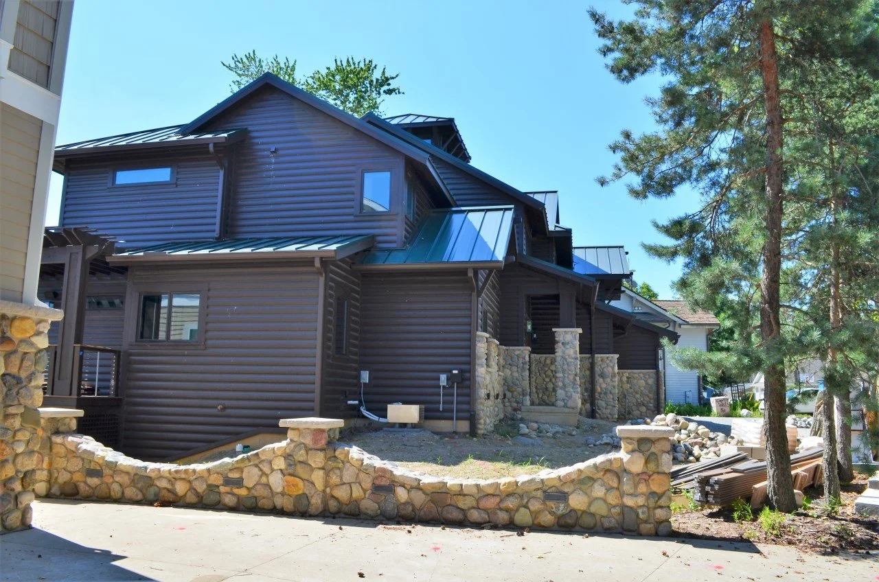 A new, dark brown house with a metal roof under construction, surrounded by large stones and trees in a residential neighborhood.