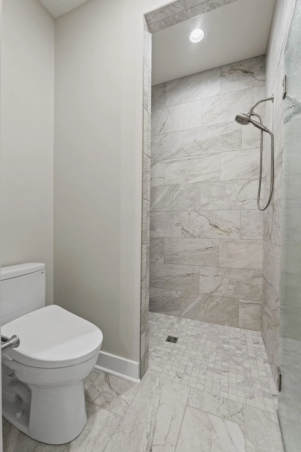 A modern bathroom with a toilet and a walk-in shower. The shower has large gray marble tiles and a handheld showerhead. There is a small window in the ceiling providing natural light.