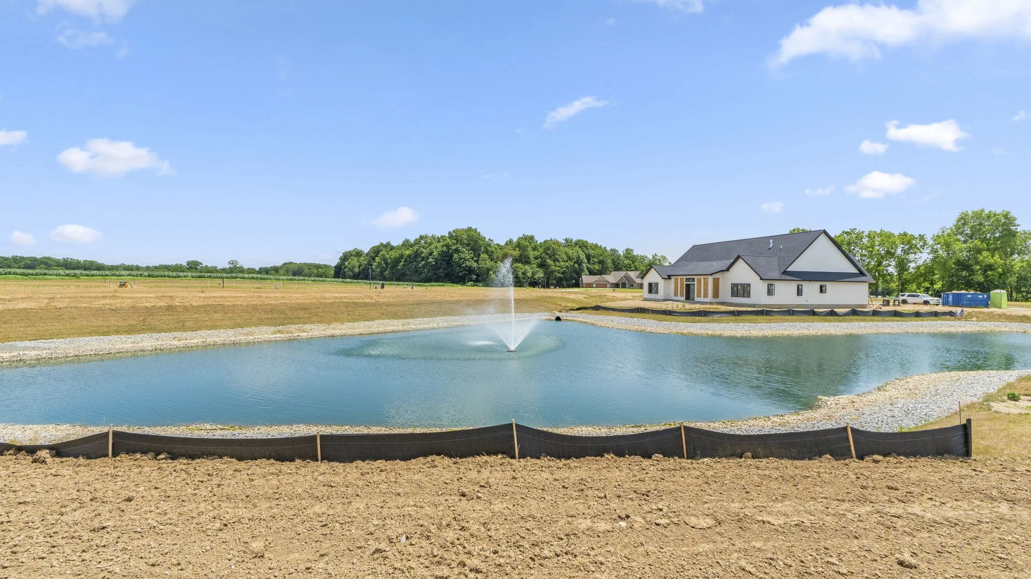 Construction site with a pond and a house in the background, blue sky with some clouds, and a fountain in the pond.