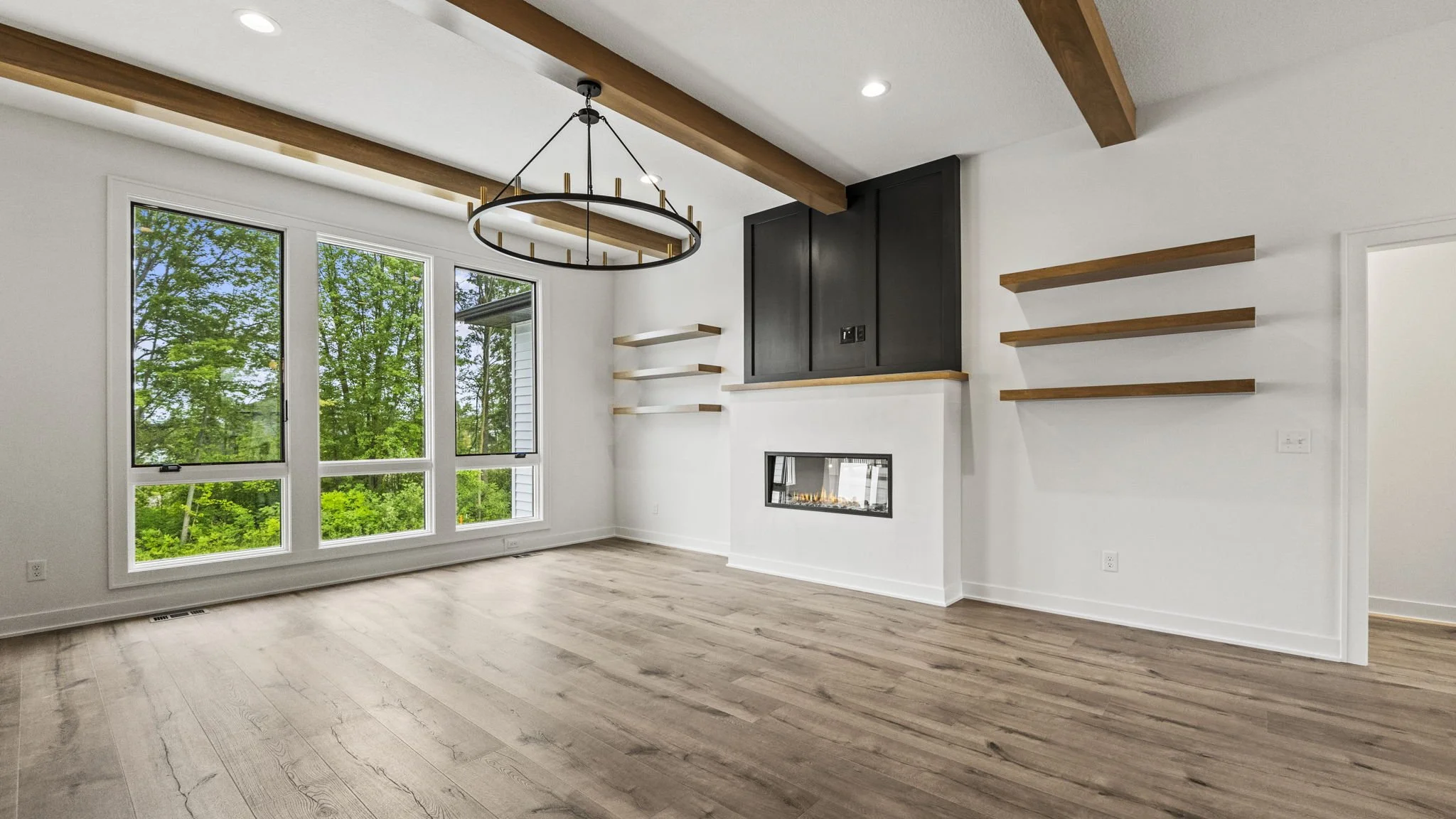 Empty living room with large windows, a modern fireplace, and floating wooden shelves on white walls, cellar ceiling with wooden beams, and a circular chandelier.