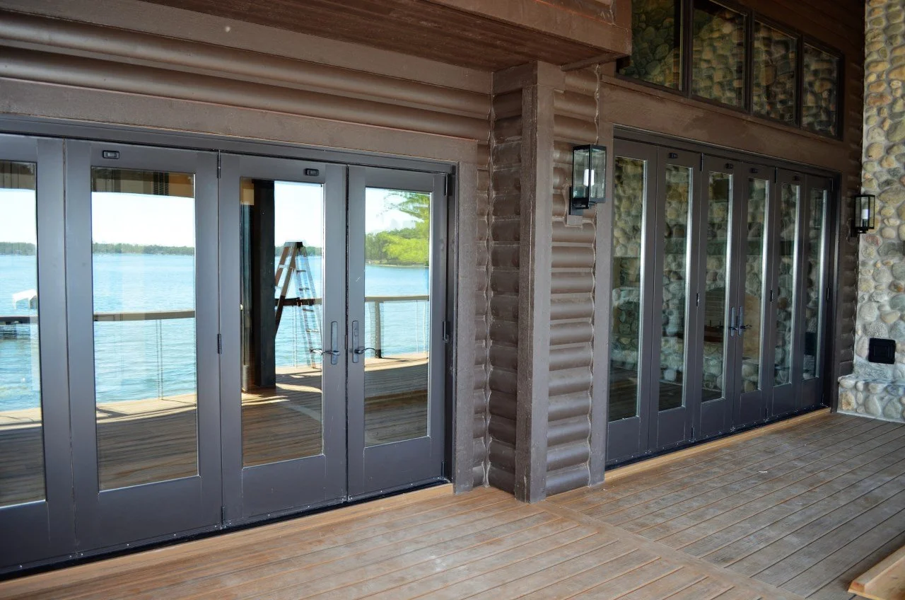 View of a waterfront porch with glass double doors and a ladder reflected in the glass, overlooking a body of water with trees in the distance.