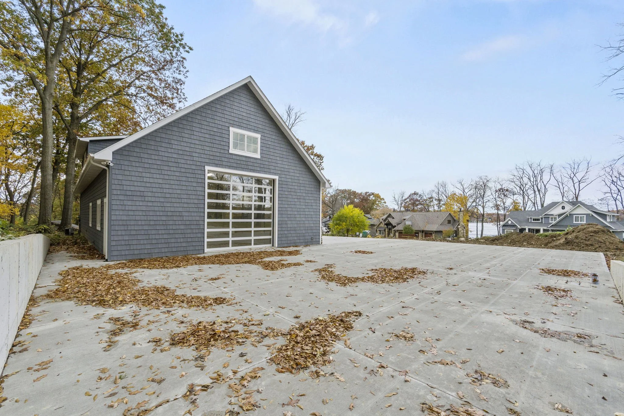 Empty driveway with fallen autumn leaves in front of a blue house with a garage, surrounded by trees with fall foliage.