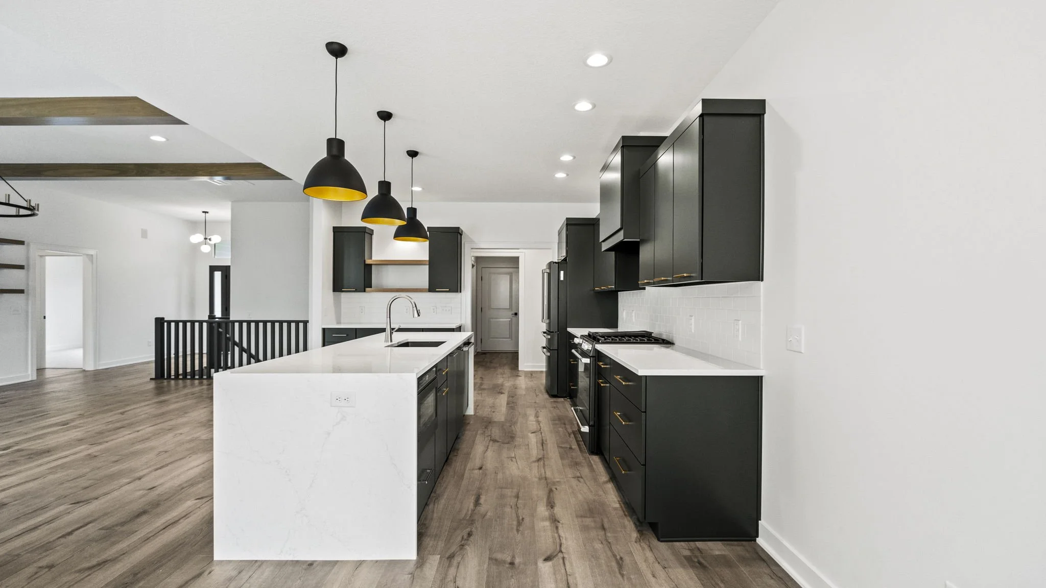 Modern kitchen with black cabinets, white countertops, a large white island with a sink, hardwood floors, black pendant lights, and stainless steel appliances.