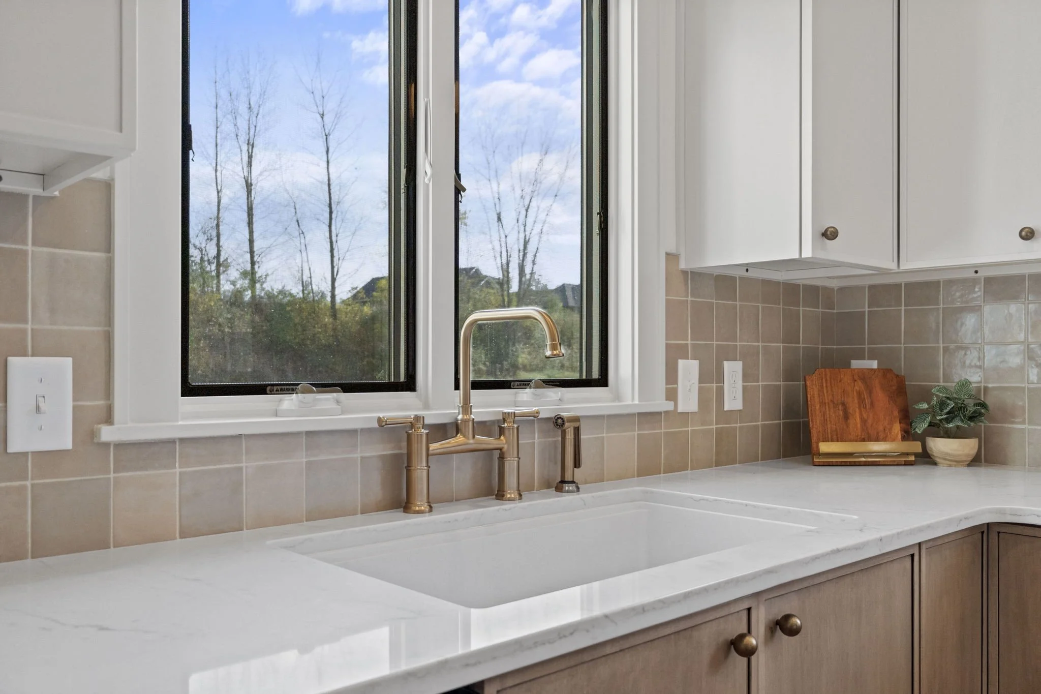 Kitchen sink with a brass faucet, beige backsplash tiles, and a window showing trees and a cloudy sky.