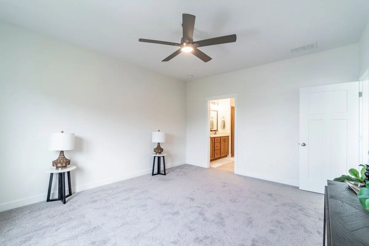 Empty bedroom with white walls, gray carpet, two bedside tables with lamps, ceiling fan, and an open door leading to a bathroom.