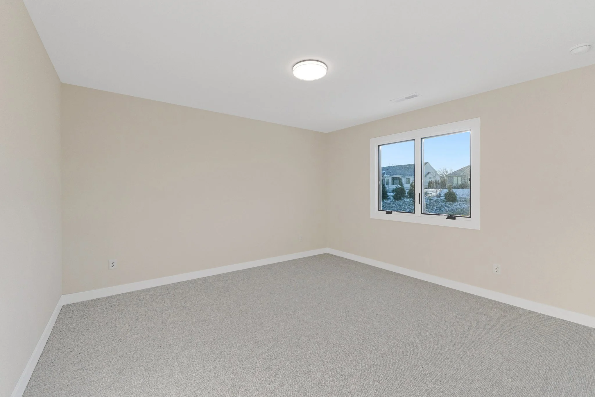 Empty beige-colored room with window overlooking neighboring houses and a white ceiling with a ceiling-mounted light fixture.