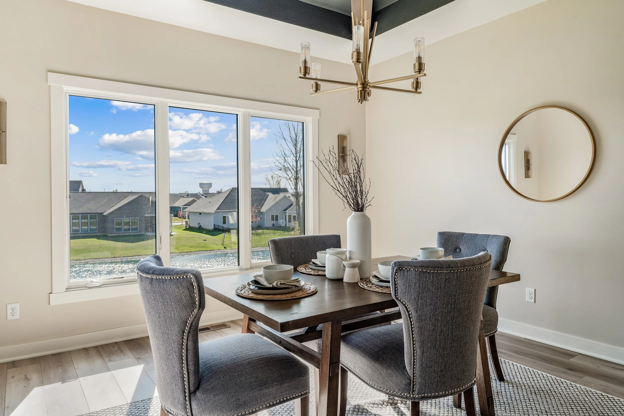 Dining room with a wooden table set with bowls, plates, and a tall white vase with branches. Four upholstered chairs surround the table. Large window showing houses, a pond, and a blue sky with clouds outside. Modern chandelier above the table and a 