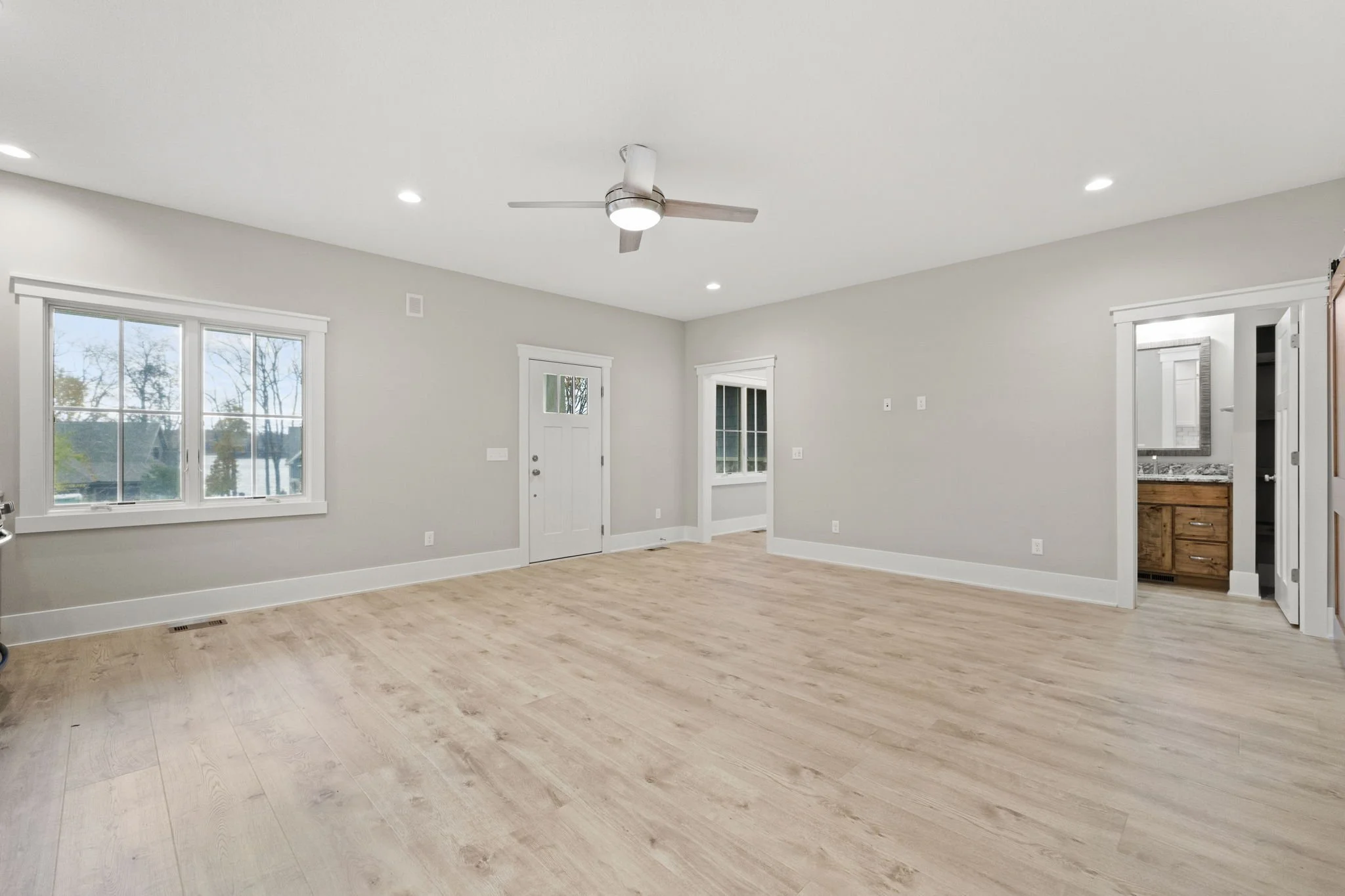 Empty living room with light wood flooring, white walls, large window, ceiling fan, and visible bathroom with wooden cabinet.